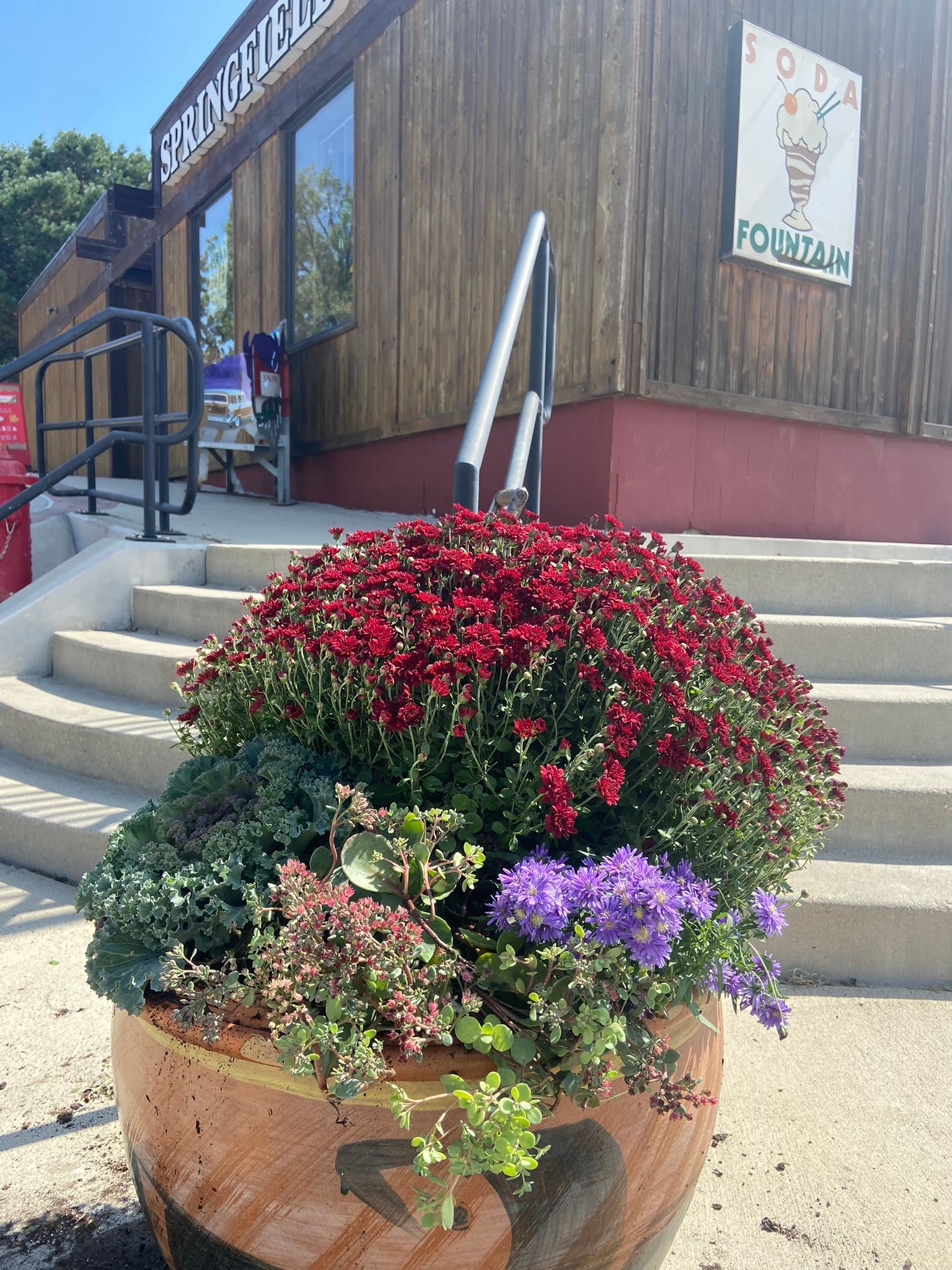 A large terracotta pot overflowing with colorful flowers, in front of a building with steps.