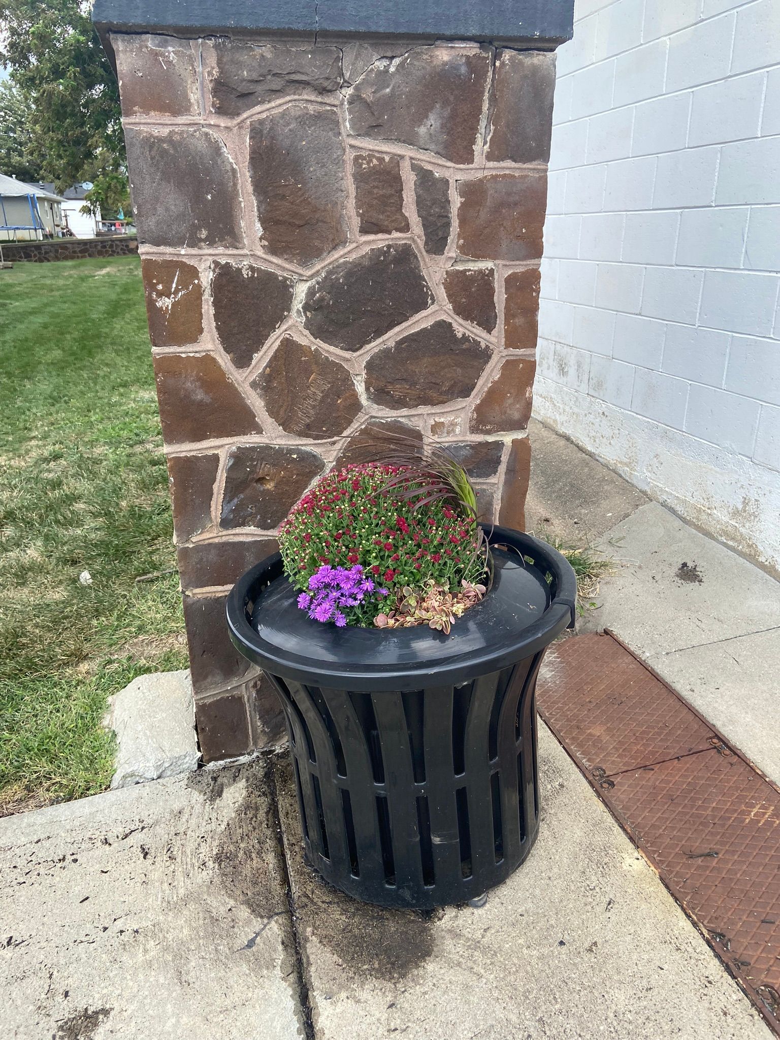 Black trash can planter with flowers in front of a stone column and a building.
