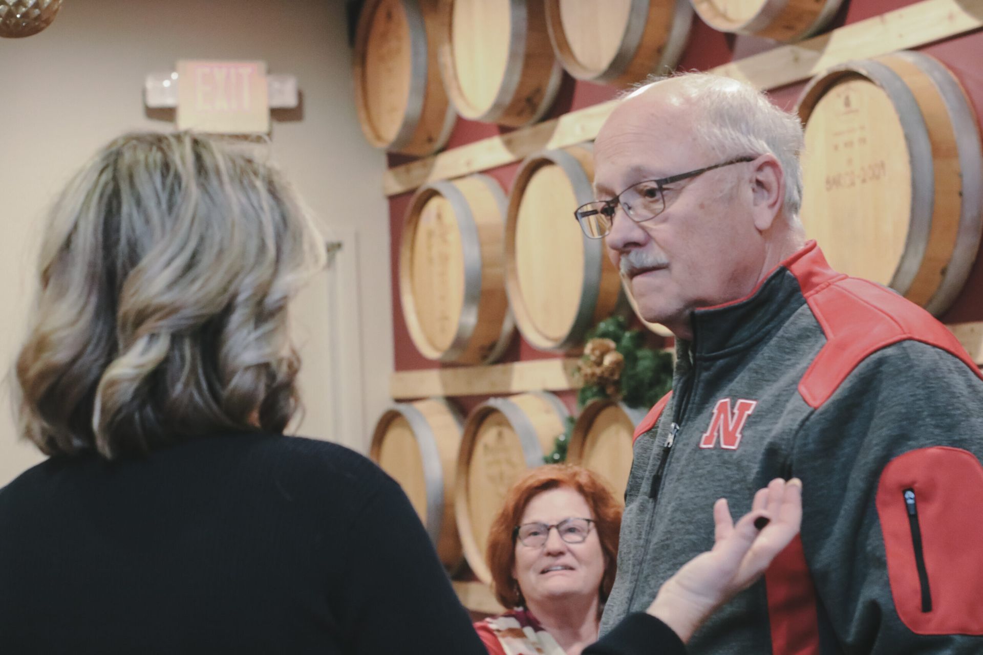 Man in red and gray jacket talking to a woman near wine barrels. Another woman smiles in the background.