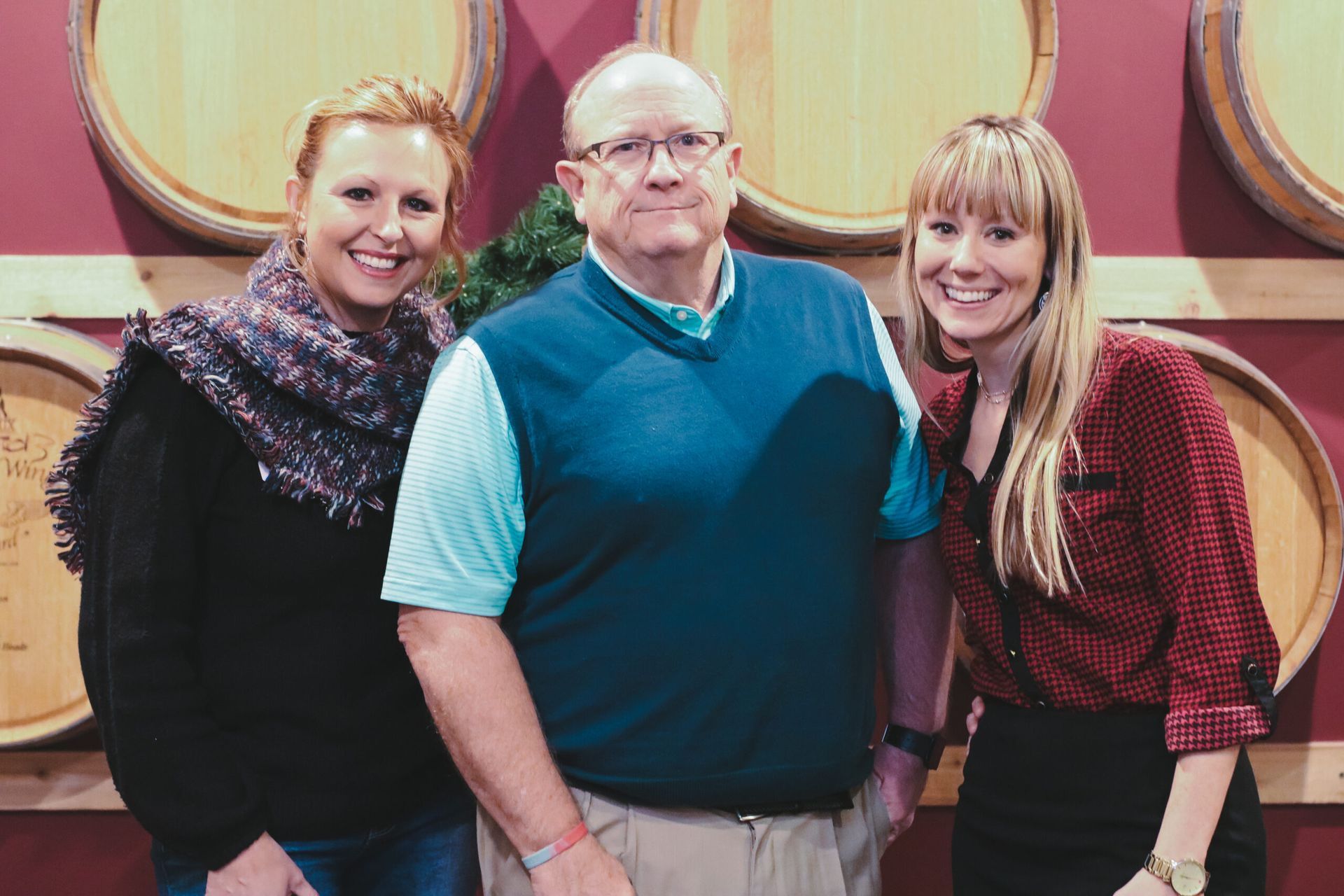 Three people smiling in front of wine barrels: woman in scarf, man in sweater, woman in red shirt.