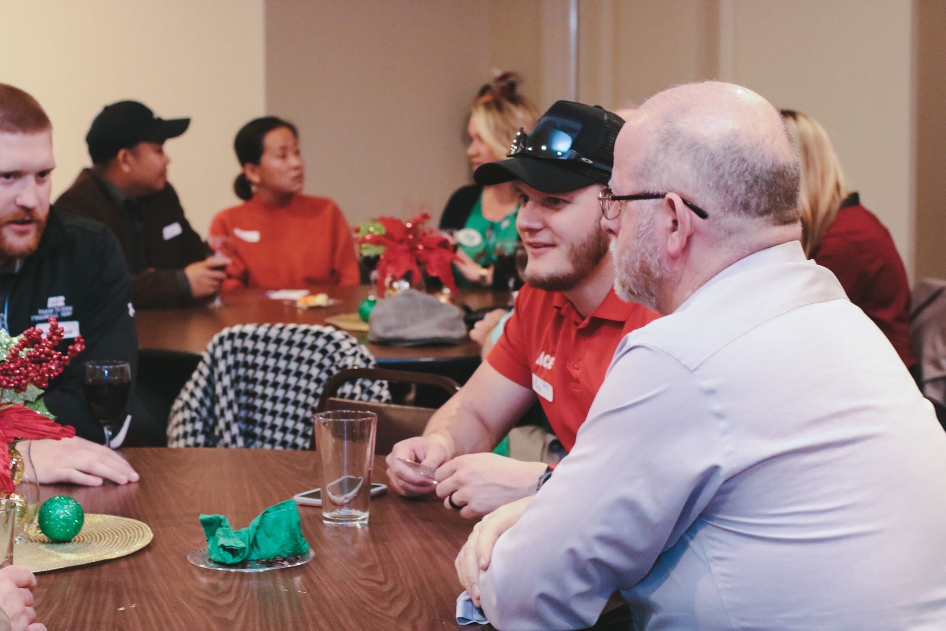 People seated at a table, conversing. Some wear red shirts. Festive decorations are visible.