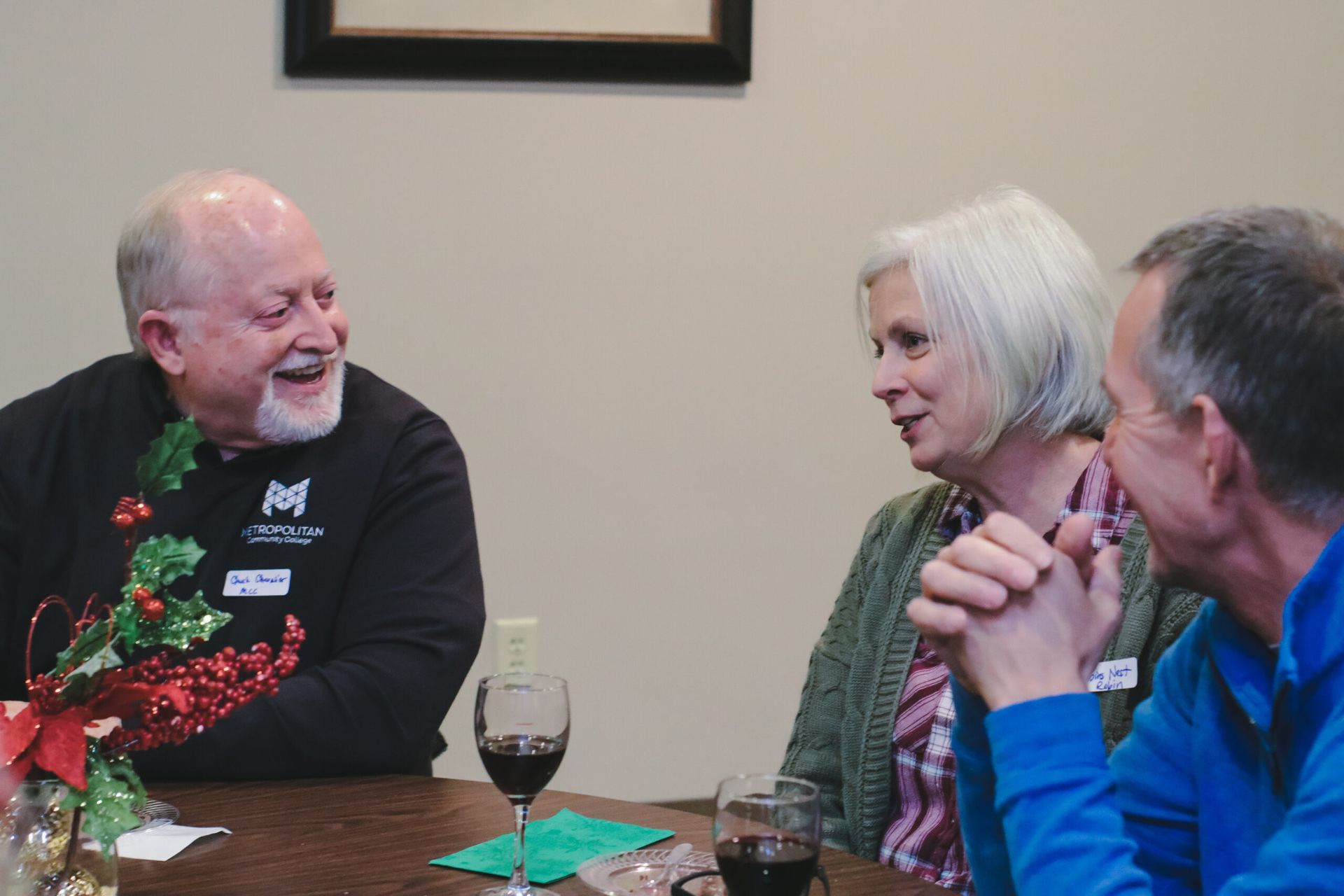 Three people conversing at a table. A man laughs, another looks on, a third person is partially visible.