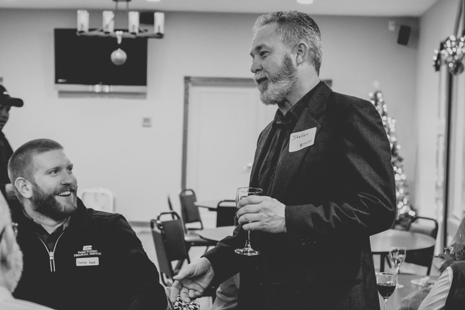 Man in blazer speaks at event, holding a glass. Another man smiles, listening. Festive setting.