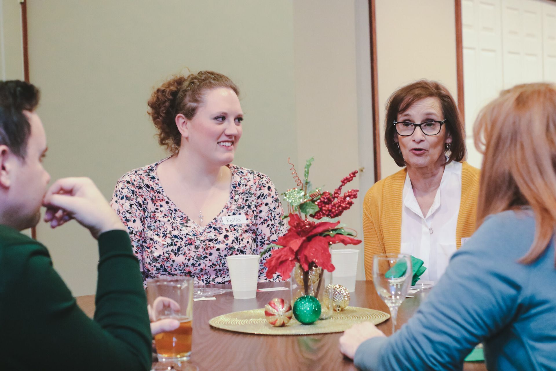 Four people at a table, two women talking. Festive centerpiece with glasses and drinks.