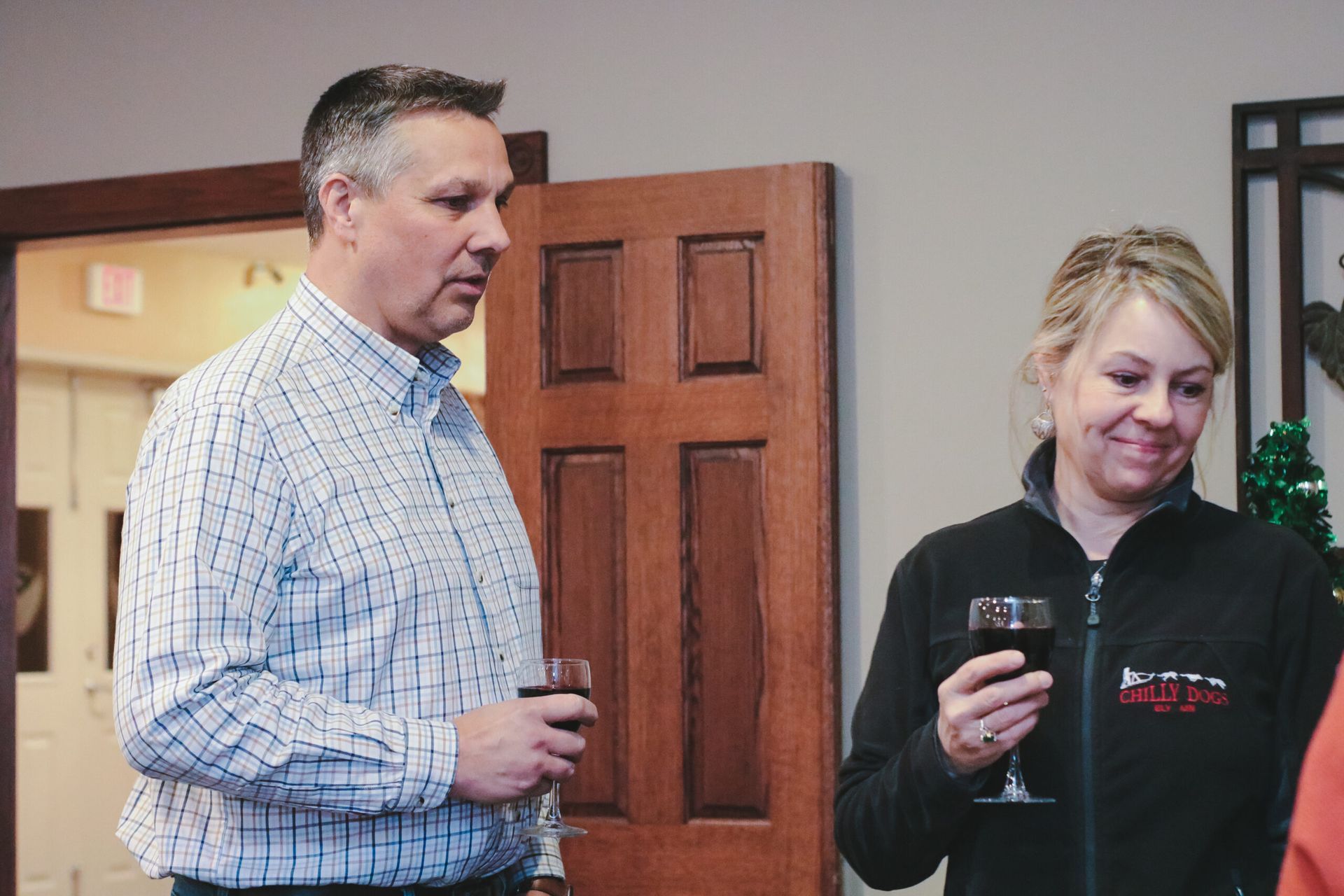 Man and woman inside, holding wine glasses, talking near a wooden door.