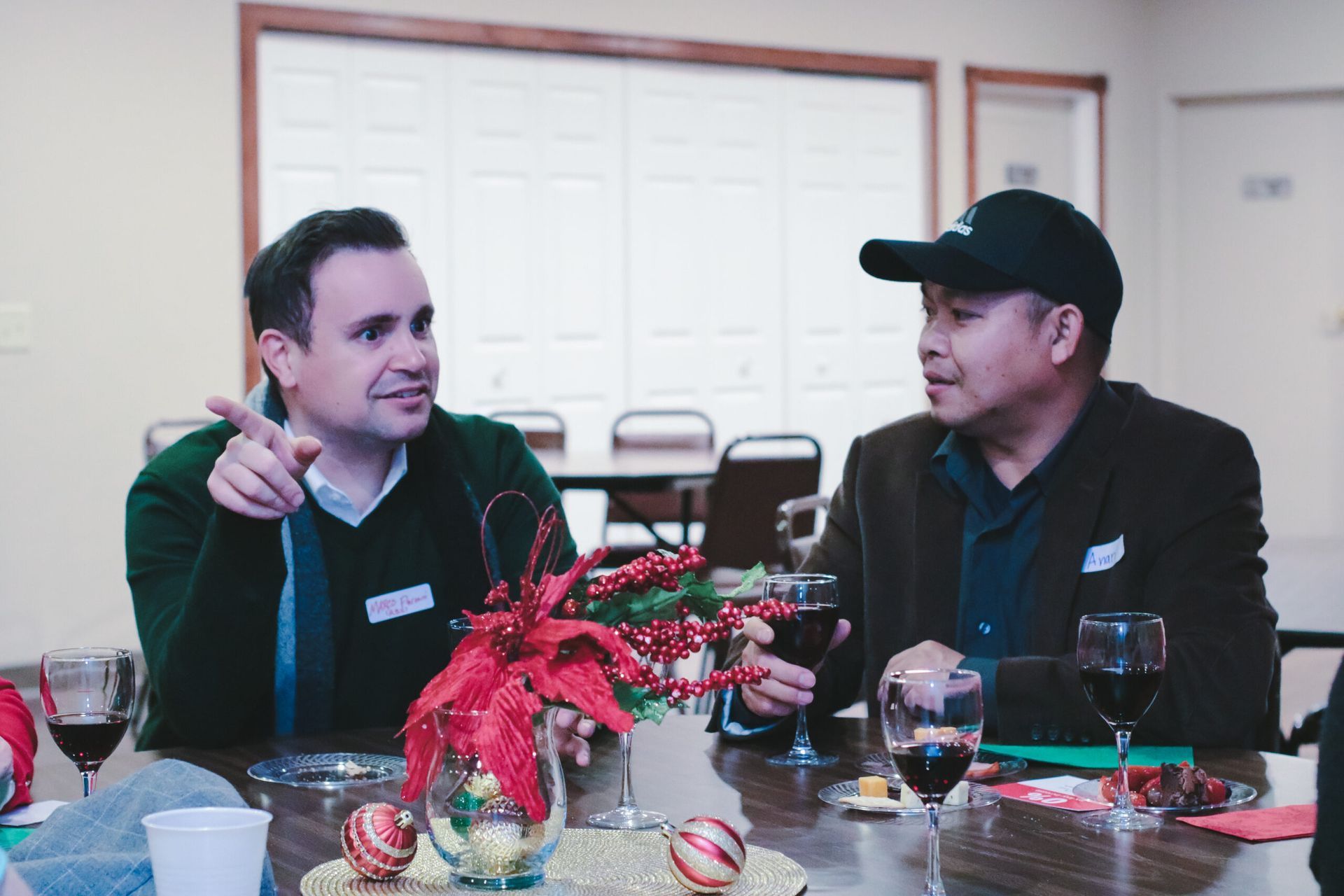 Two men seated at a table, talking and holding wine glasses. Festive centerpiece in foreground.