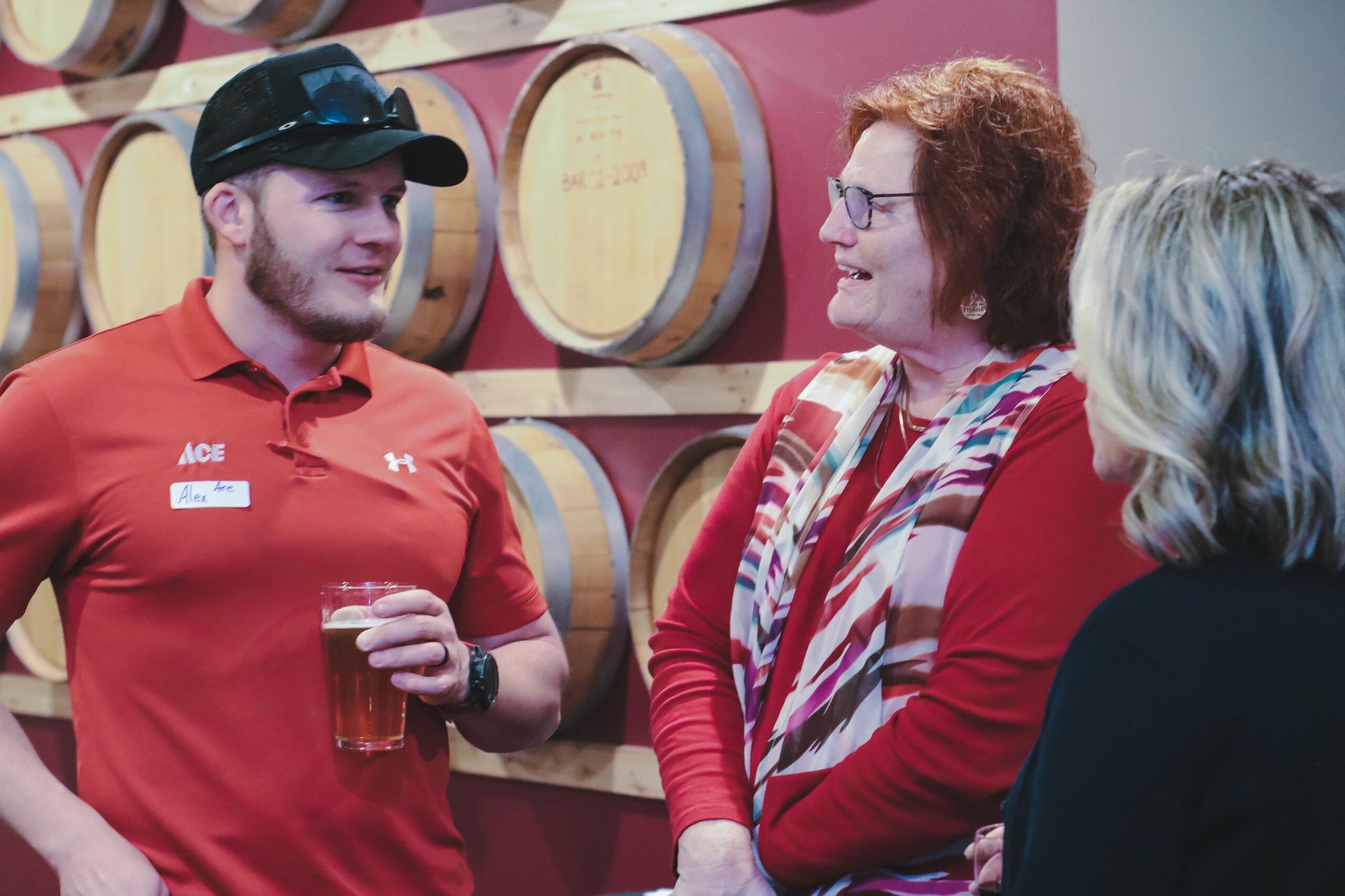 Man in red shirt talks to two women in front of wooden barrels.