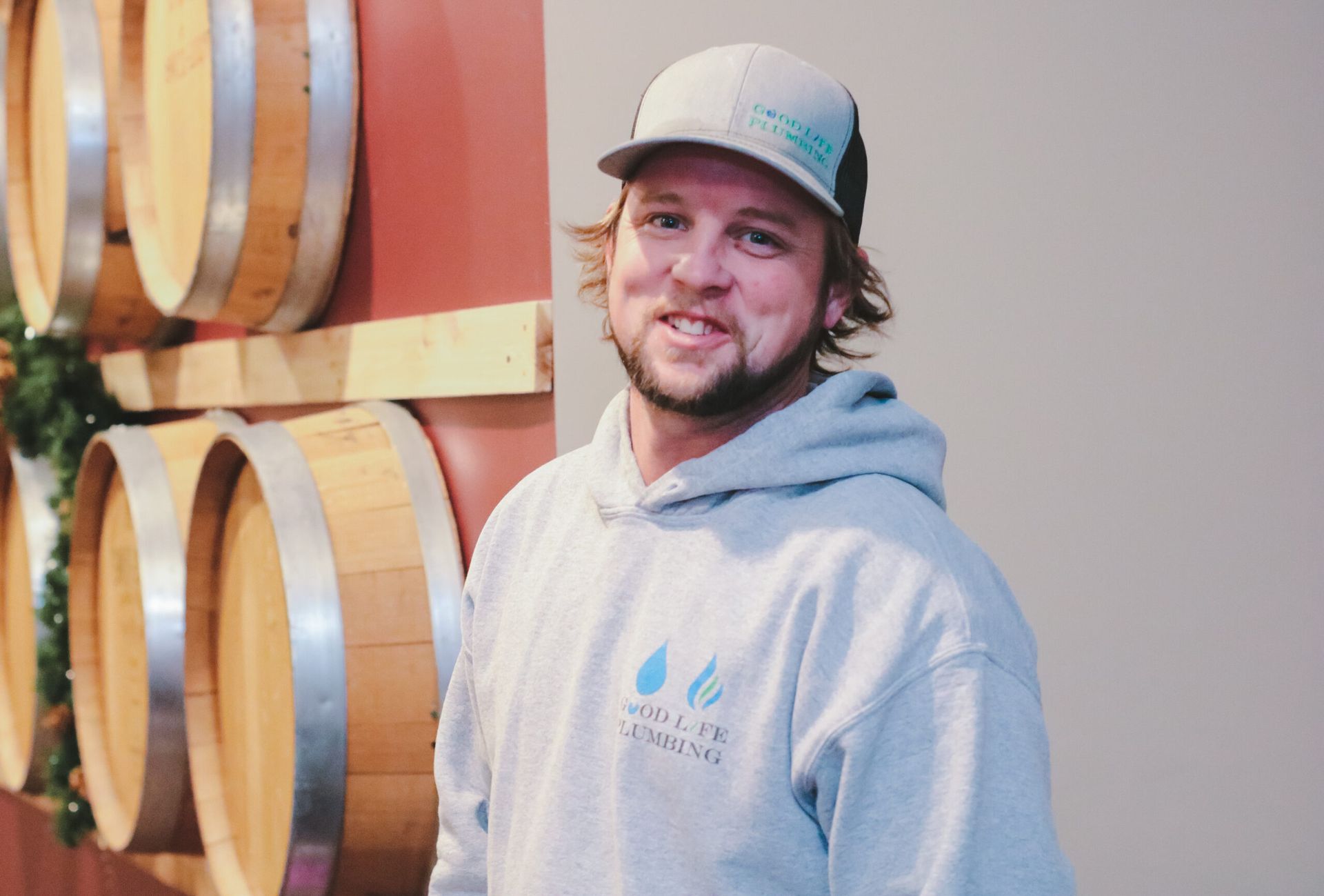 Man in grey hoodie and cap smiles in front of wine barrels.
