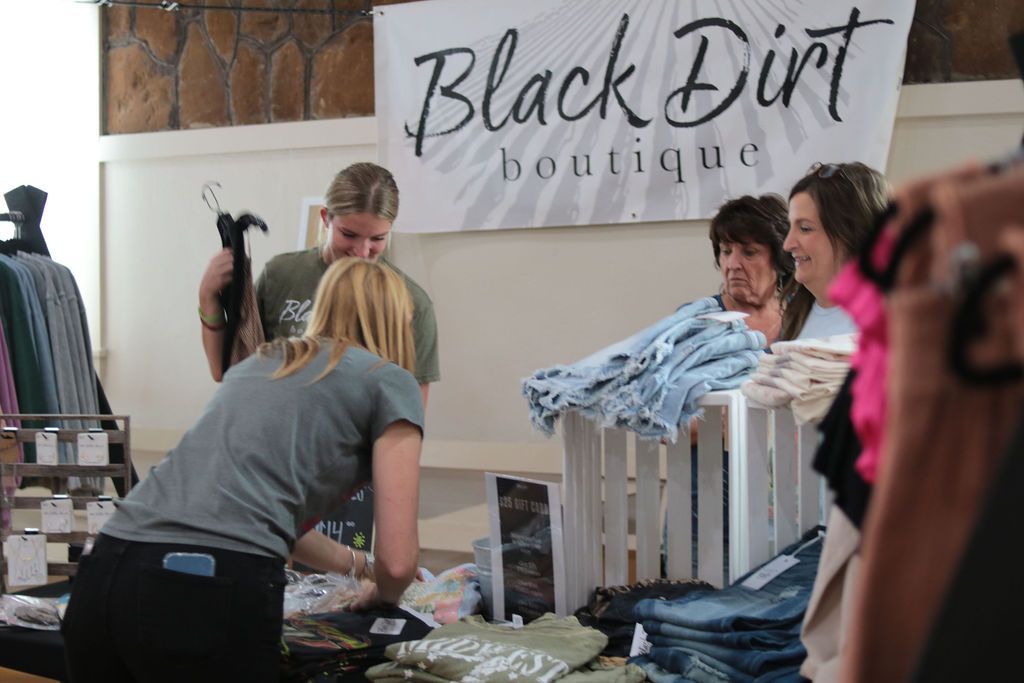 Black Dirt Boutique shop, customers and staff. Woman behind table holds items, smiling. White sign: Black Dirt Boutique.