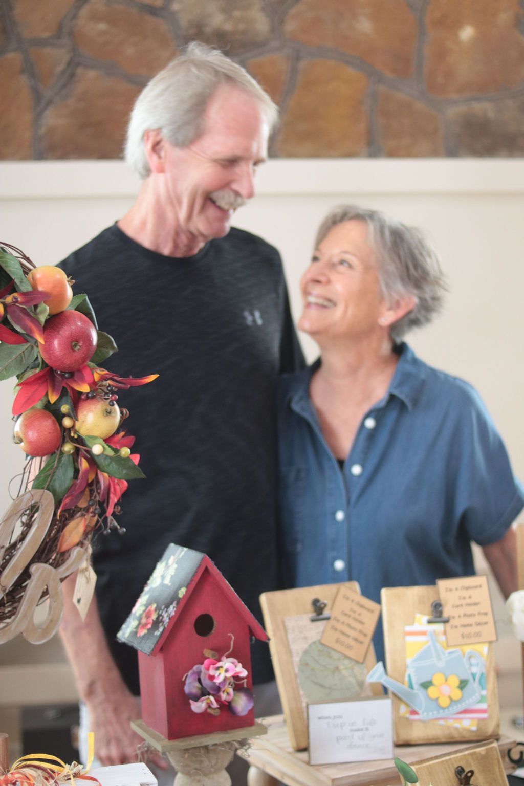 Smiling couple standing behind a table of crafts, including a birdhouse and gift bags.