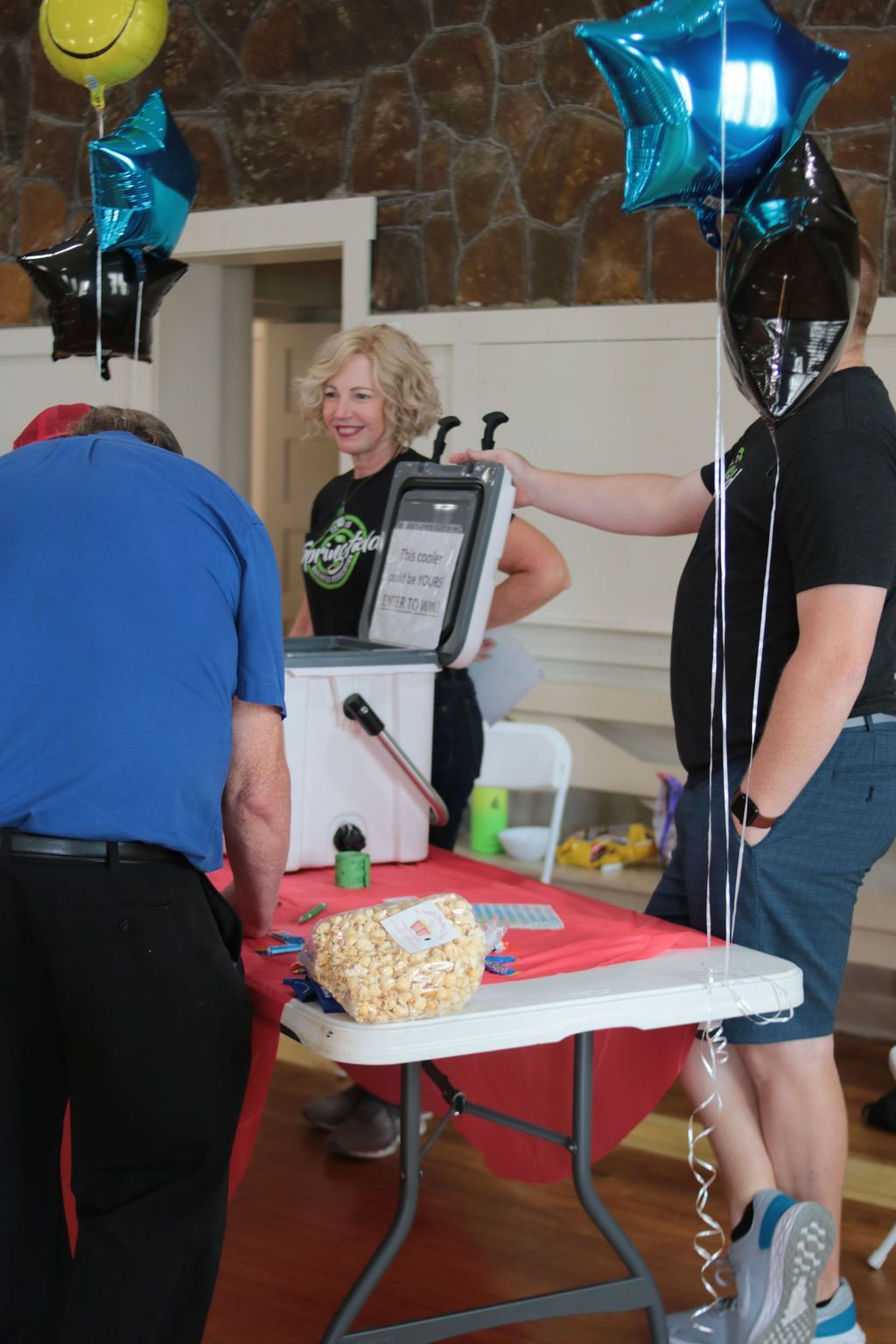 People at a table with balloons. Woman smiling, holding a cooler. Table has popcorn and sign.