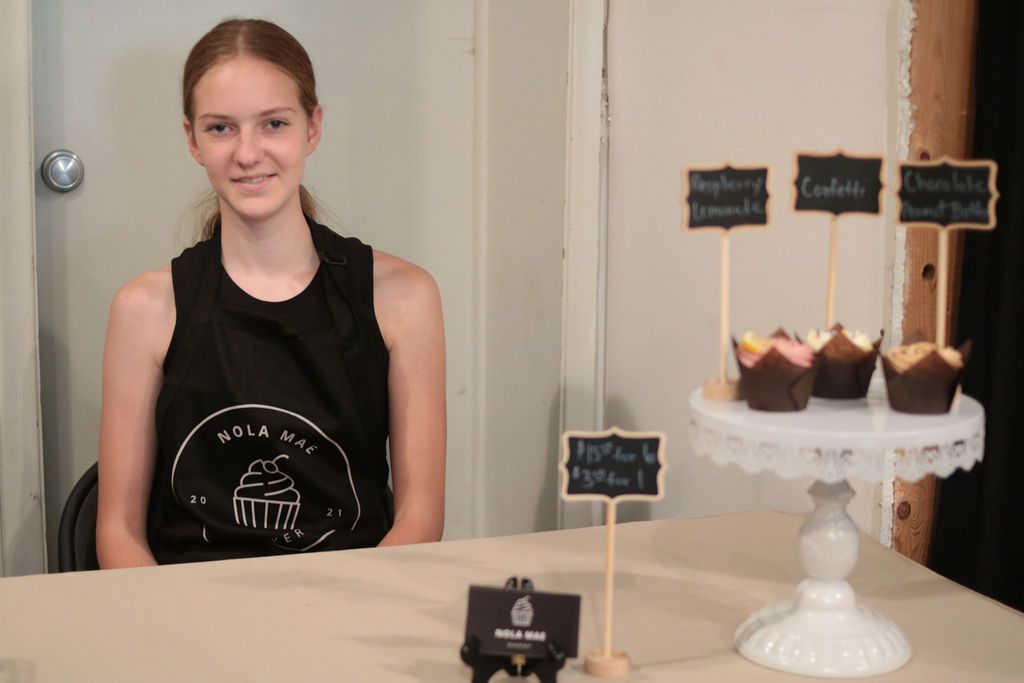 Woman seated at table with cupcakes on a stand. Signs identify flavors.