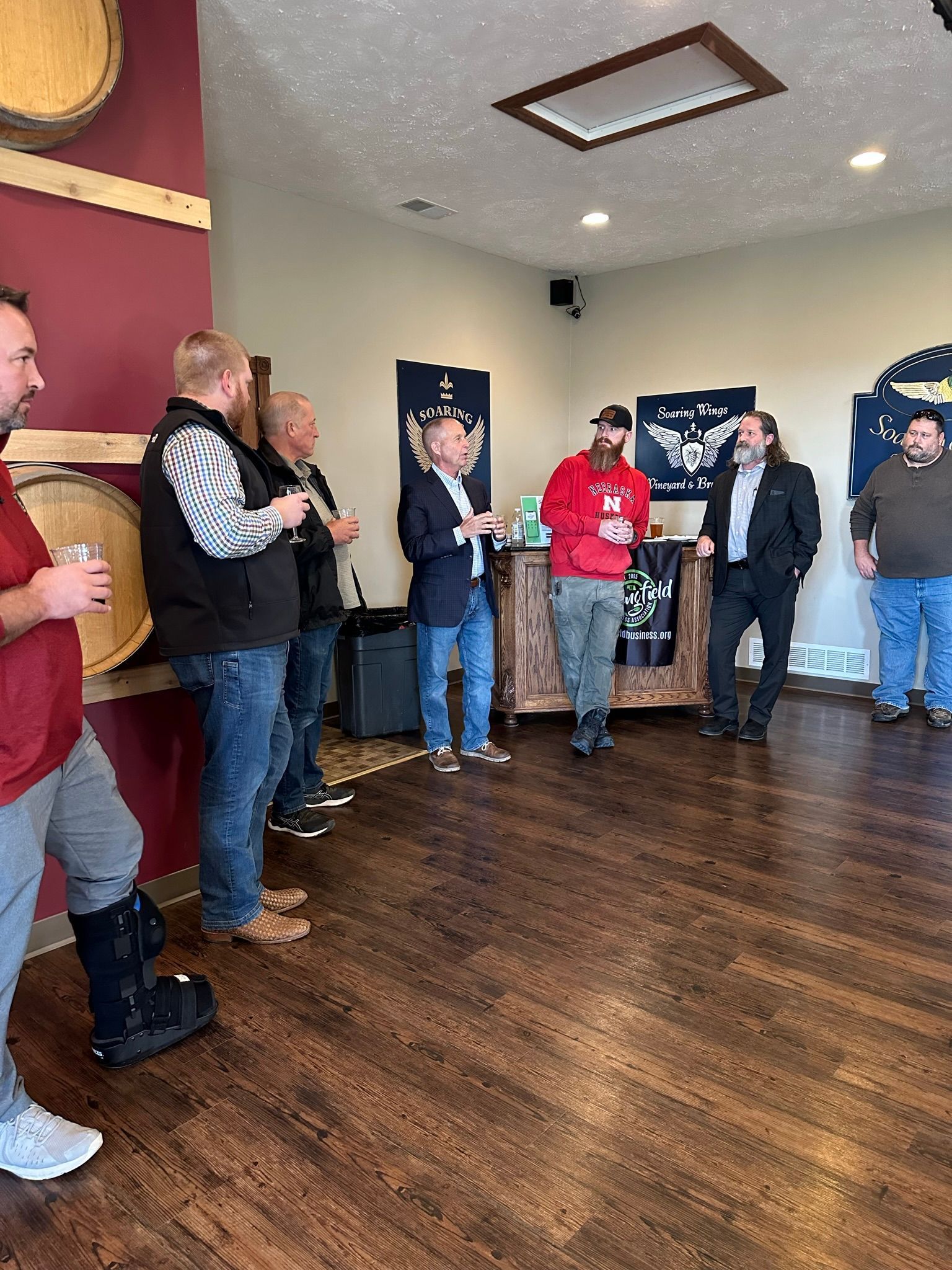 People gathered in a room with wood floors. Some are speaking, others listening. Barrels and decorative items are in the 