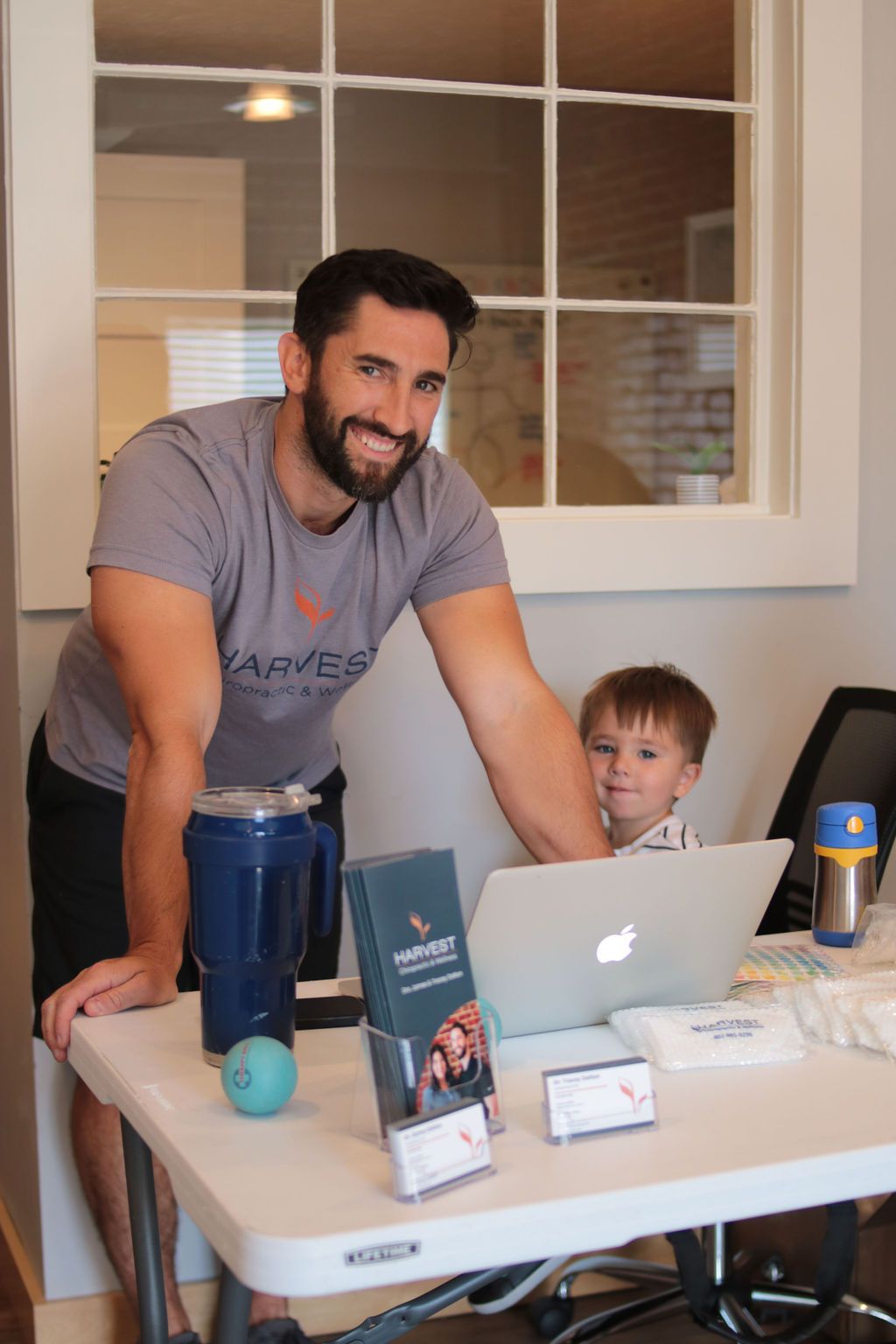 Man smiles near laptop with child, at a white table. Harvest branding is present, along with a blue cooler and a window.