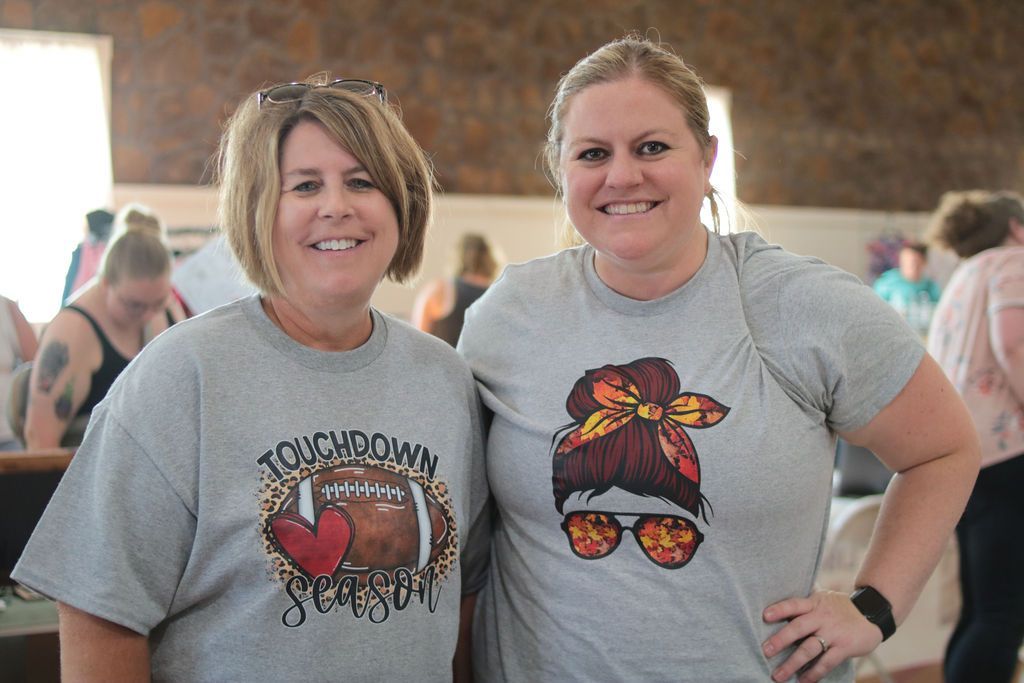 Two women in gray t-shirts smiling, one with a football design, the other with a messy bun.