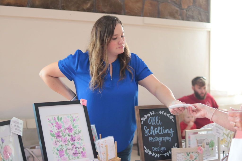 Woman in blue shirt at a booth, handing item to someone. Watercolor artwork and sign are on display.