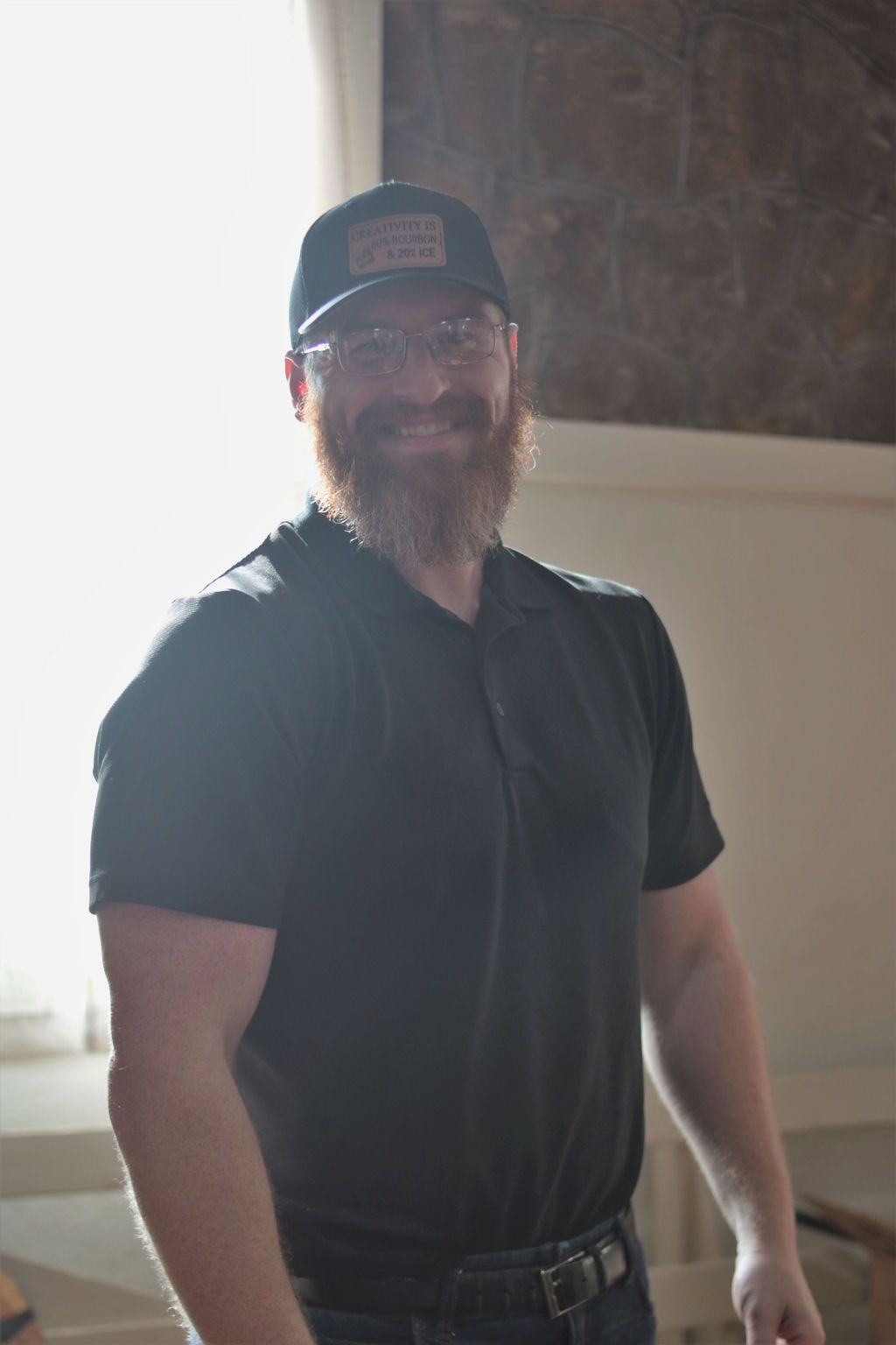 Man in black shirt and baseball cap smiles, standing near a window.