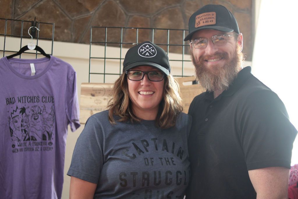 Two people wearing hats, smiling. Clothing items on display behind them.
