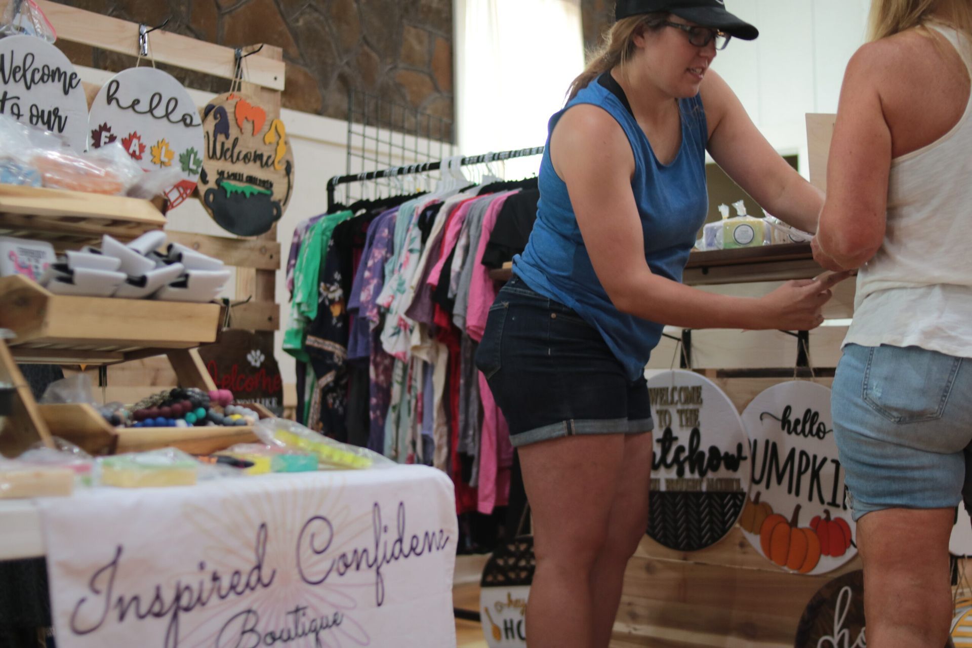 Woman helping customer at a boutique stand, with clothing rack and signage.