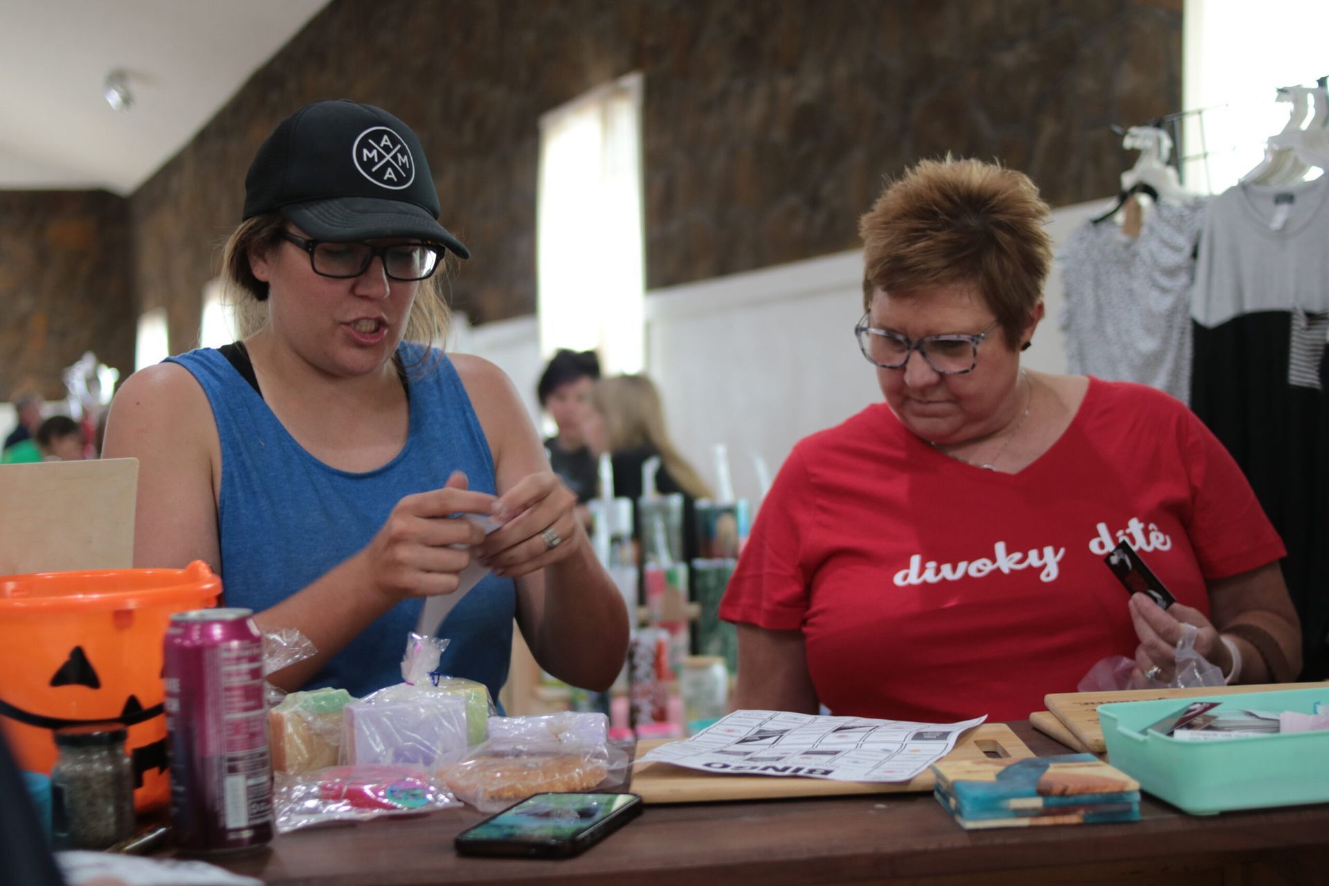 Two women at a table. One in blue tank top and hat works on something. Other in red shirt looks on, both wearing glasses.