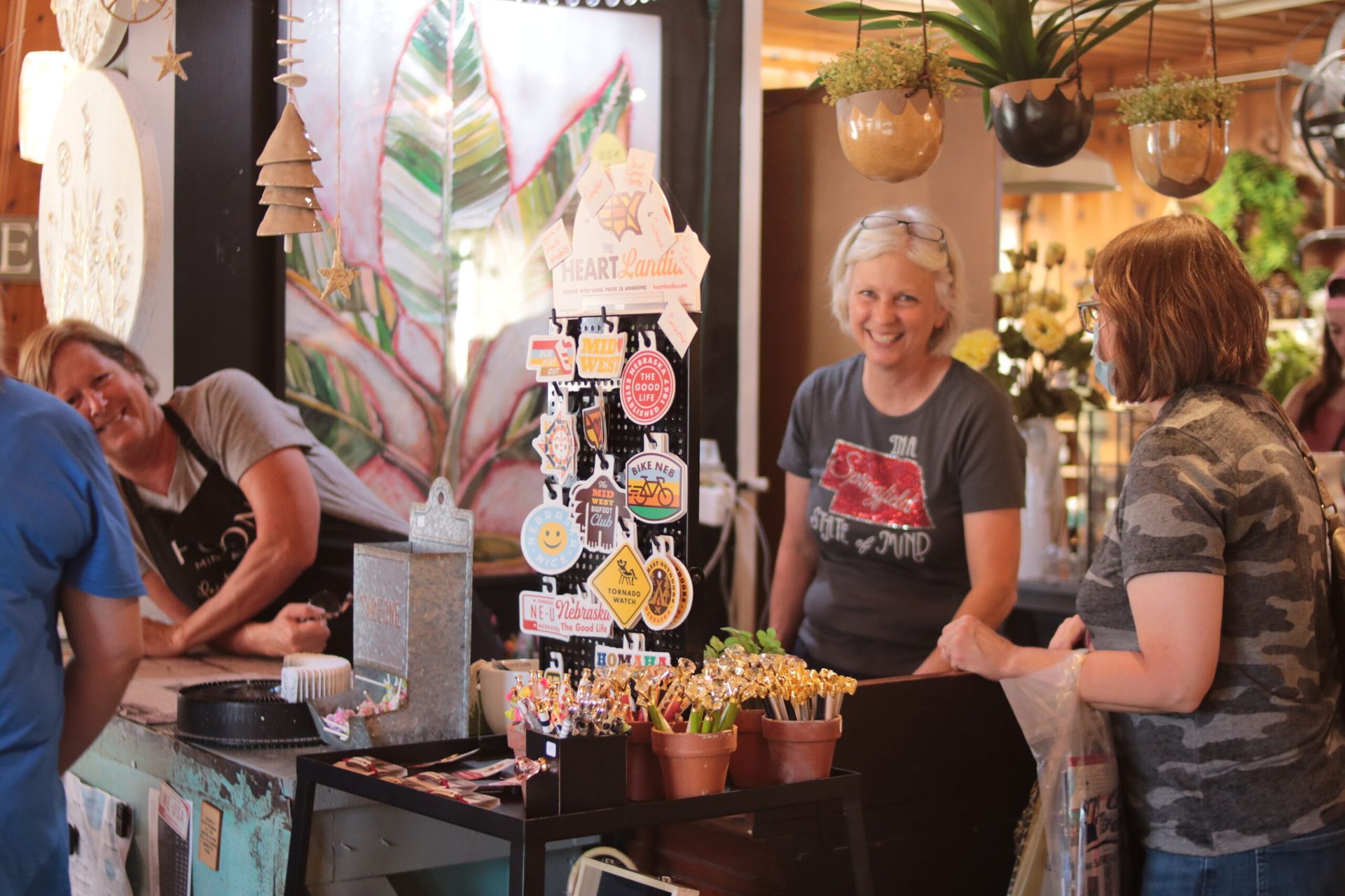 Two women at a shop counter, one smiling at a customer. Plants and merchandise displayed; indoor setting.