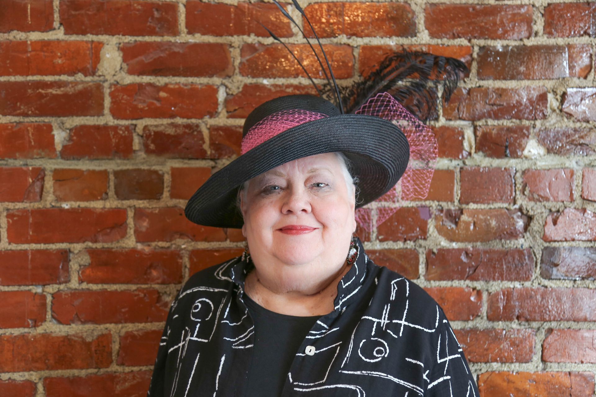 Woman in hat, black and white jacket, in front of a brick wall.