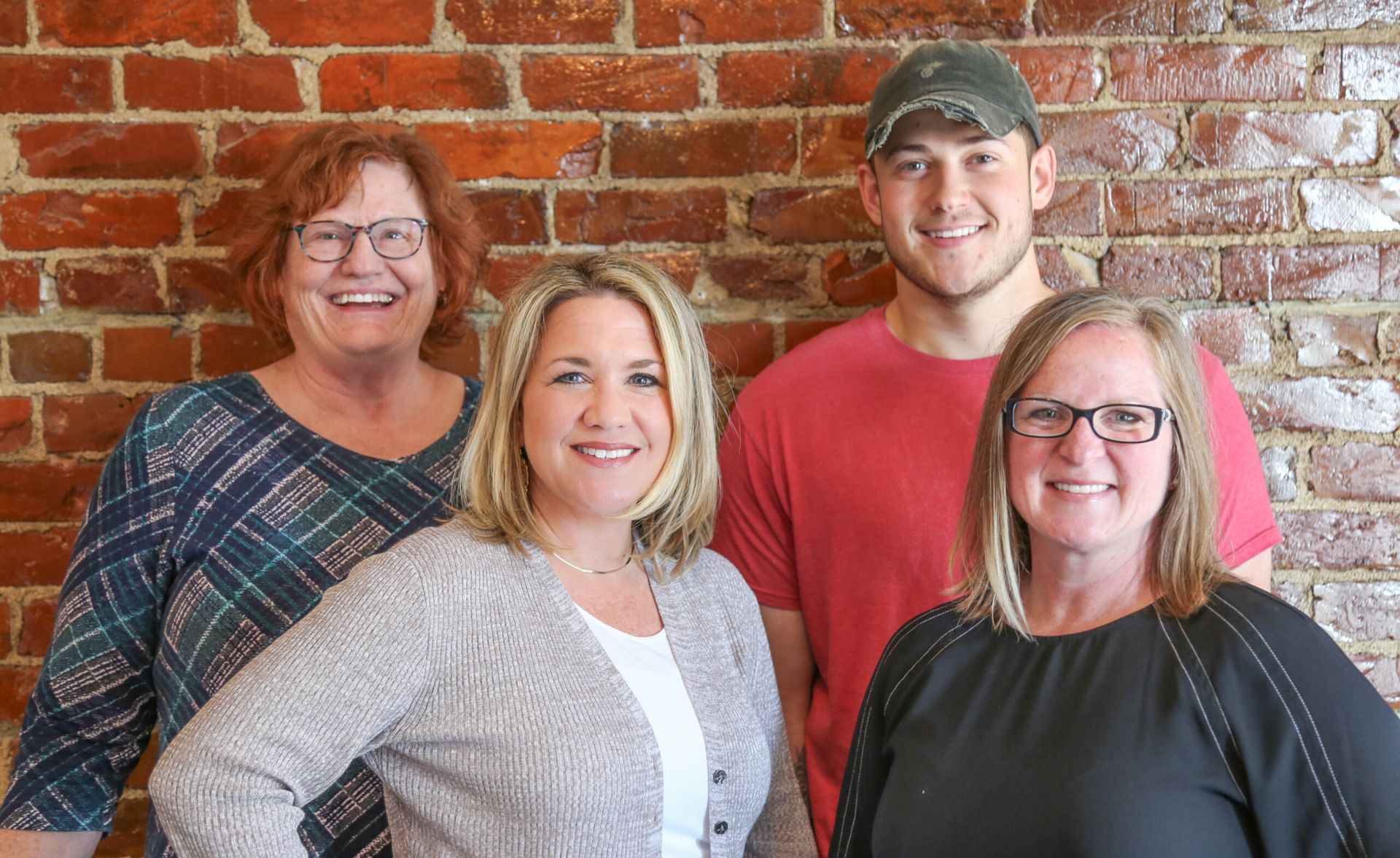 Four people smiling in front of a brick wall. Two women in glasses, one with a green plaid top. Man in a cap and red shirt.