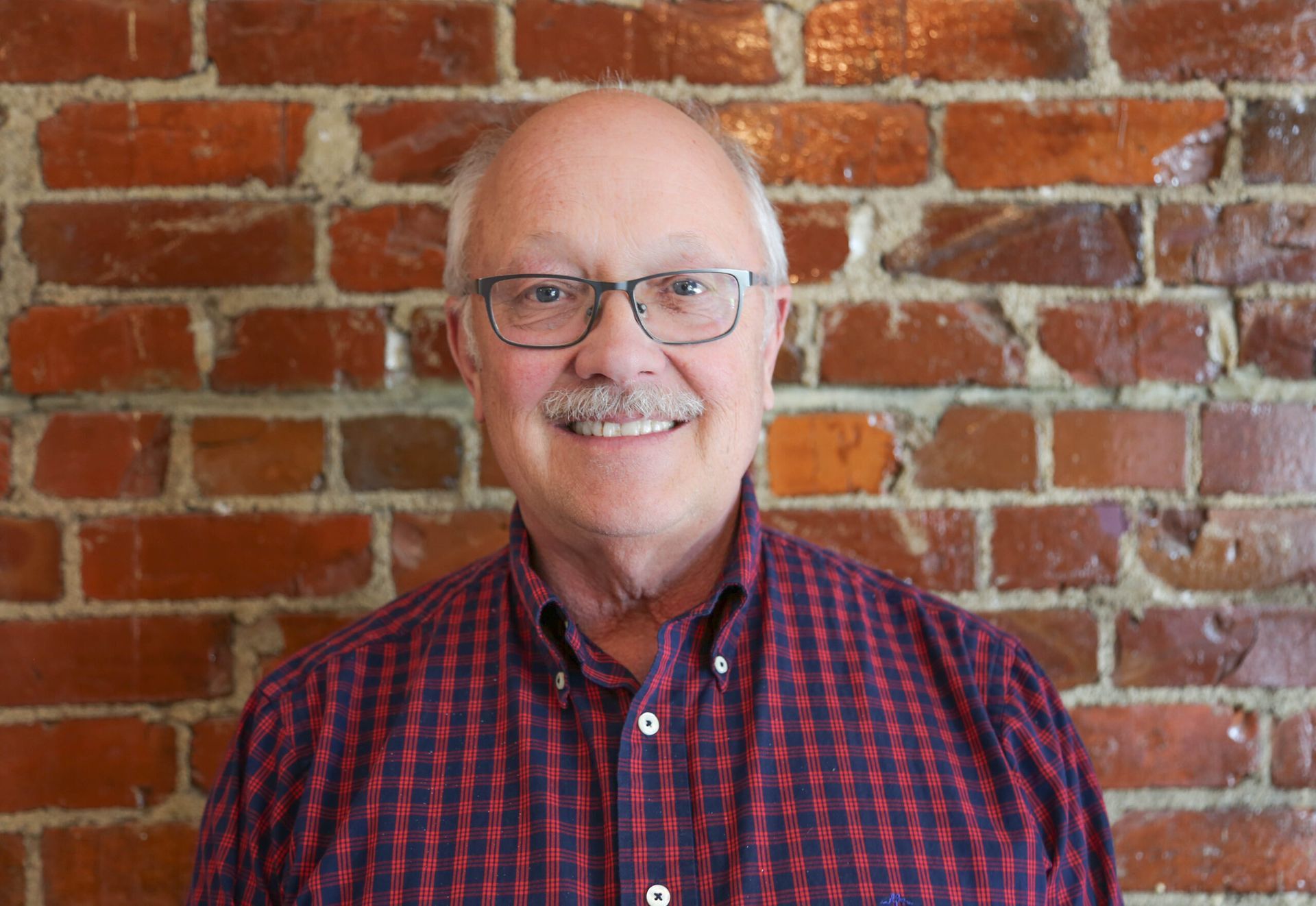 Man with glasses and mustache smiles in front of a brick wall, wearing a red and black checkered shirt.