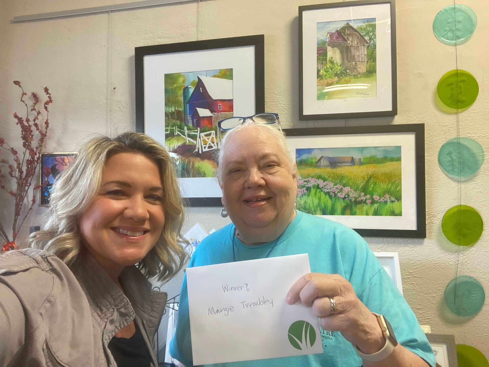 Two smiling women pose with artwork, one holding an envelope, in an art gallery.