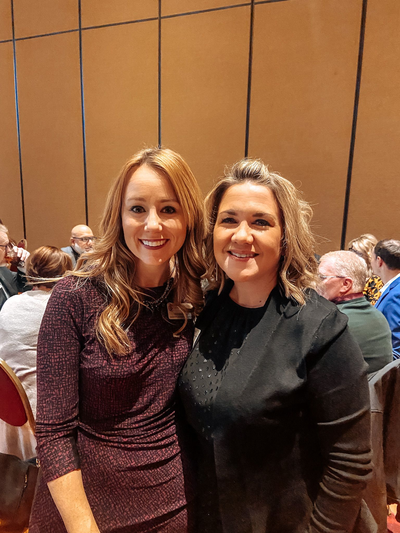 Two women smiling, posing for photo at an event. One wears a dark dress, the other a black top. People in background.