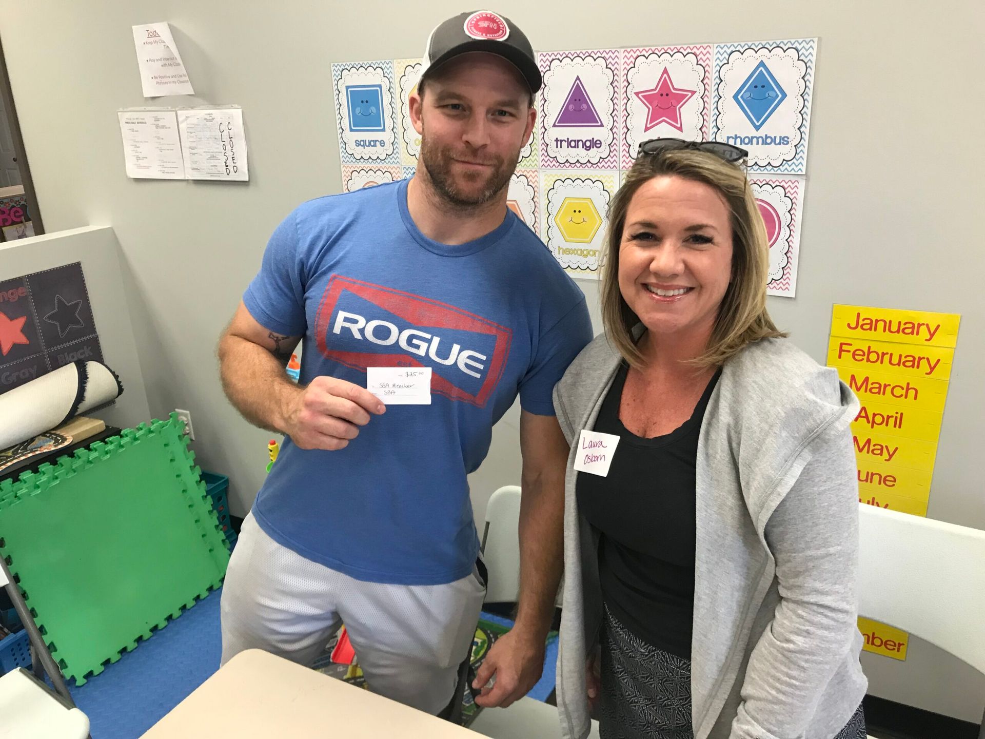 Man in blue shirt and woman in grey cardigan smiling, holding a card, inside a classroom.