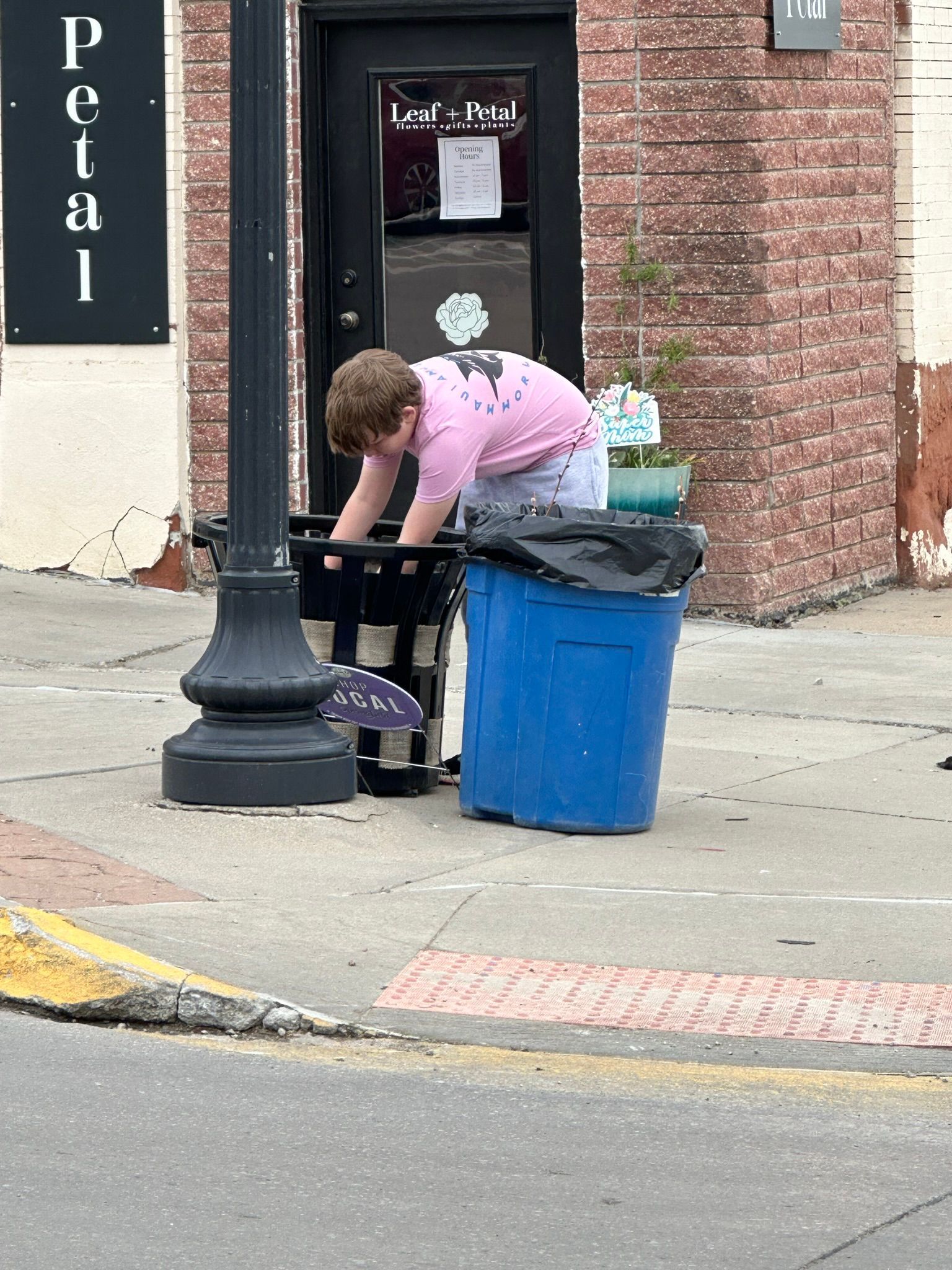 Person in pink shirt reaching into a trash can next to a black lamp post and blue trash bin on a sidewalk.