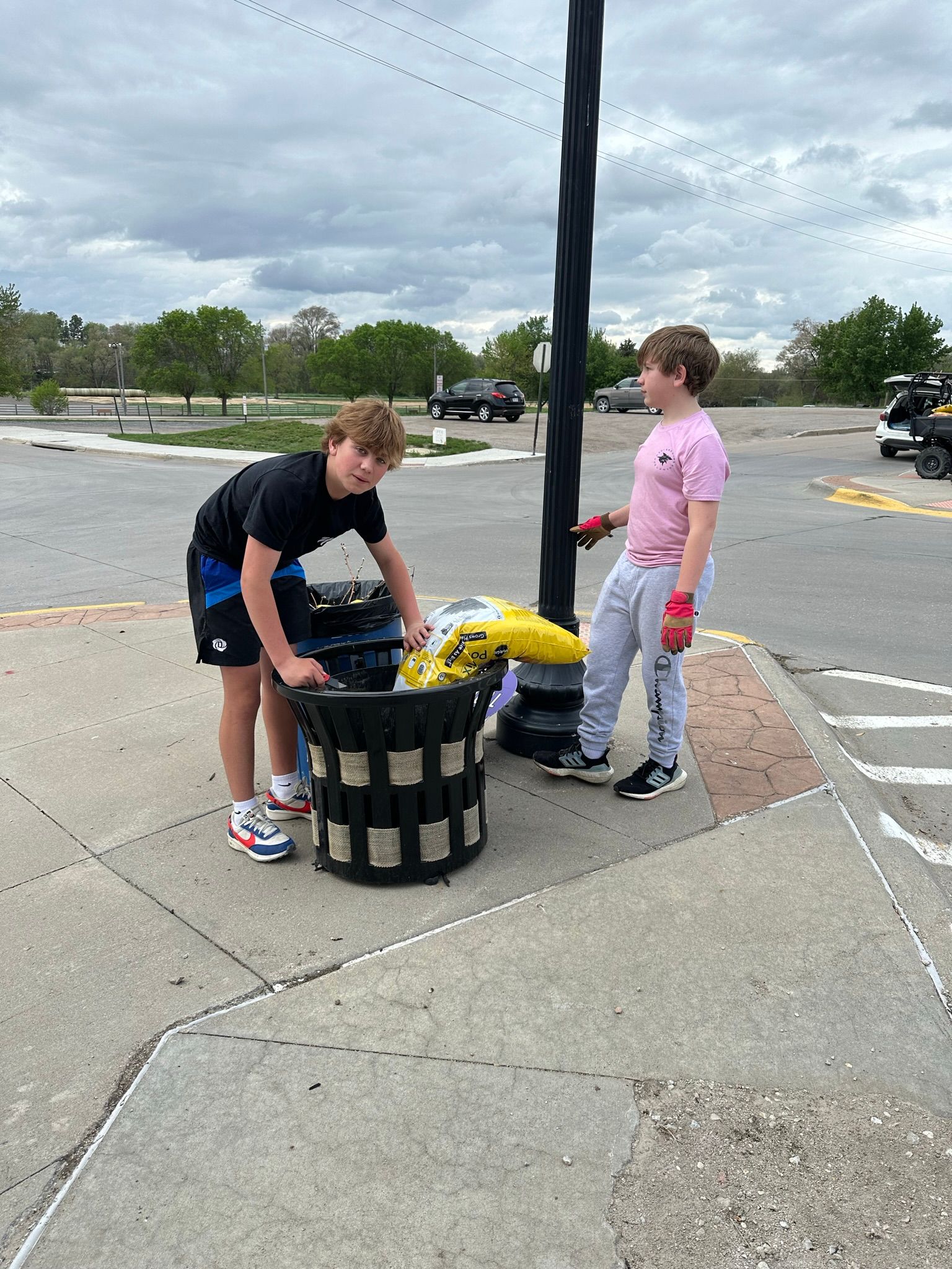 Two people putting trash in a large outdoor bin near a parking area.
