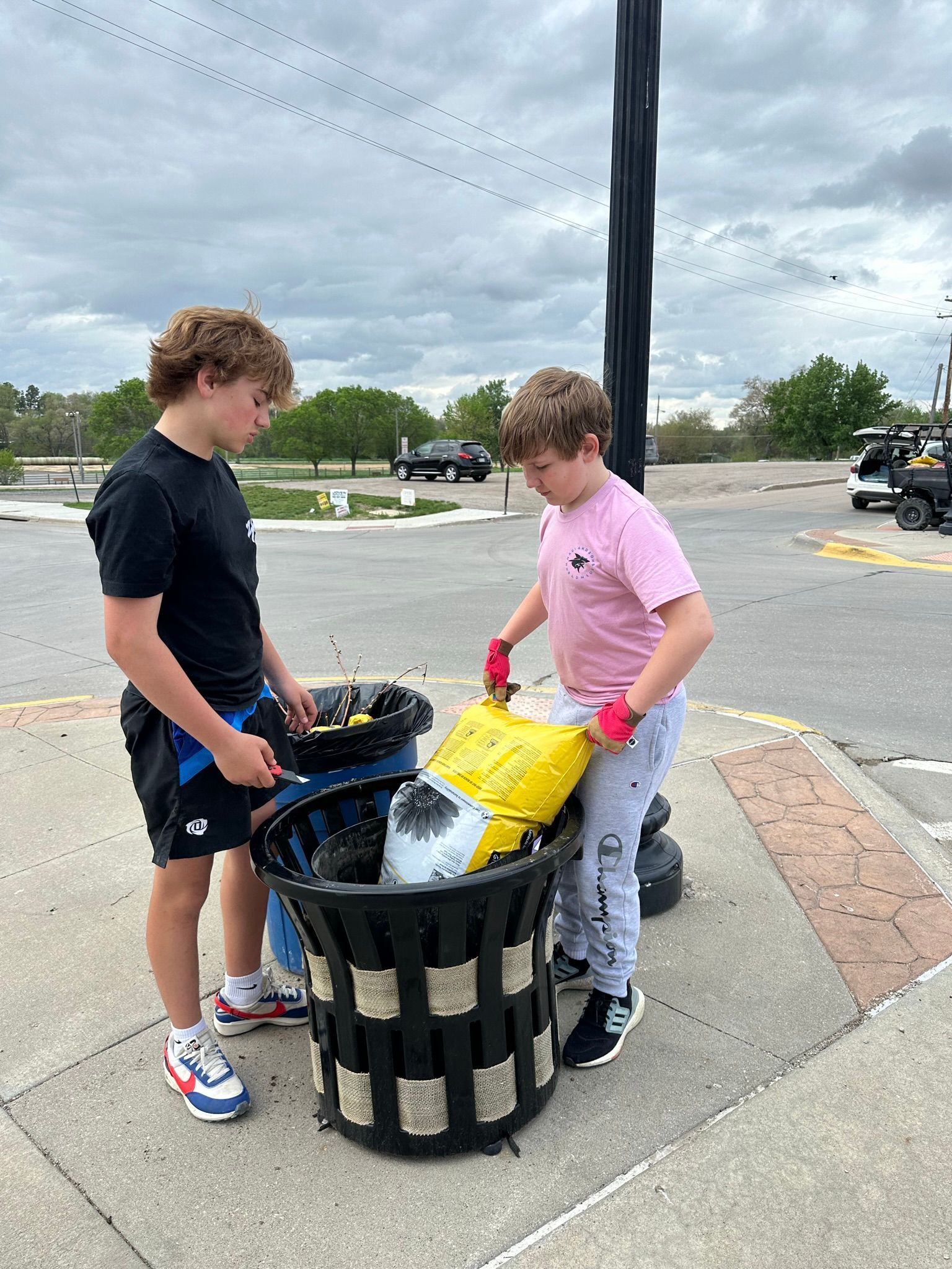 Two boys putting trash into a large, outdoor trash can on a cloudy day.