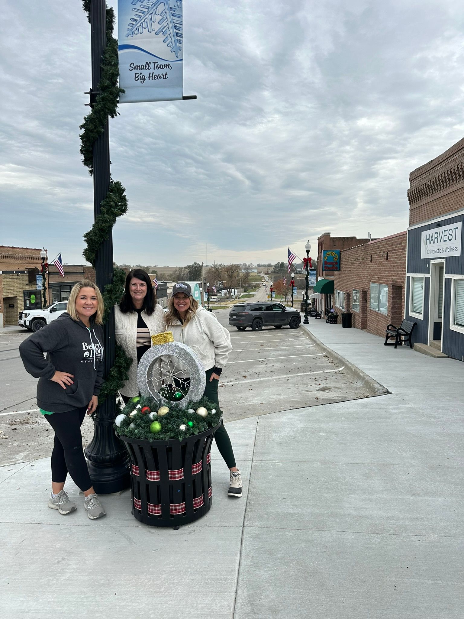 Three people pose by a decorated planter, street scene with shops, gray sky.