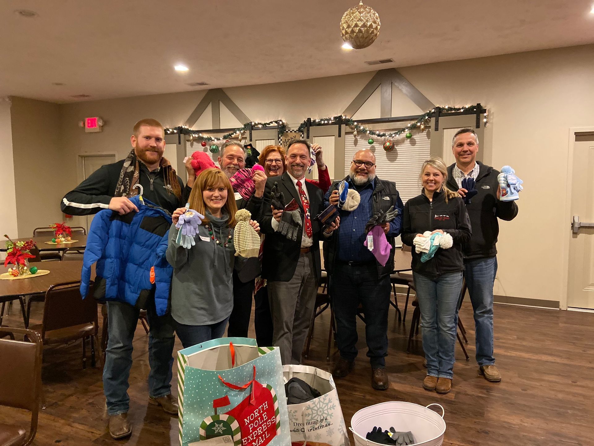 Group of people holding up winter clothing, smiling, inside a decorated room.