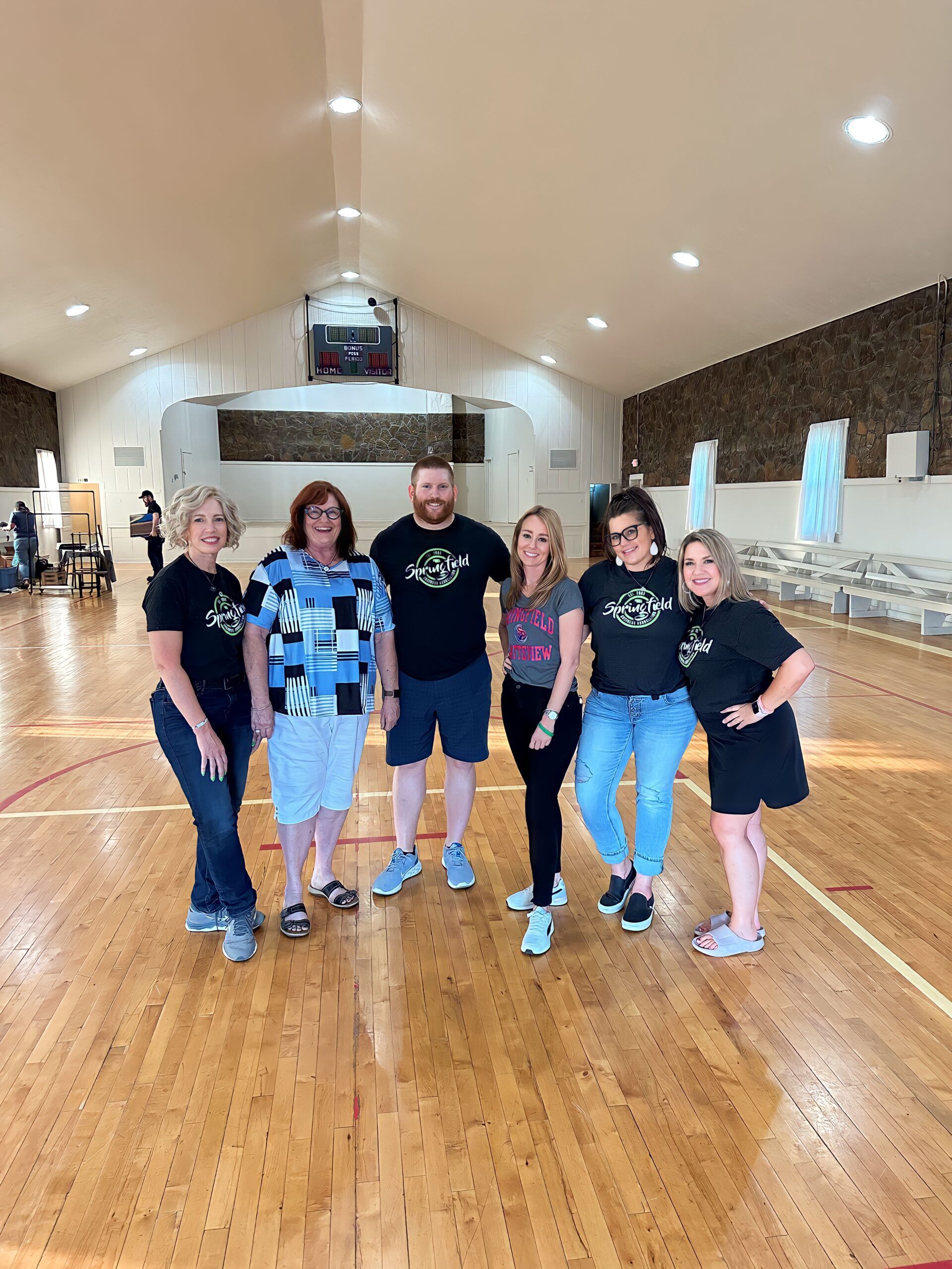 Group of people posing in a gymnasium. Six adults wearing casual attire stand on a wood floor.
