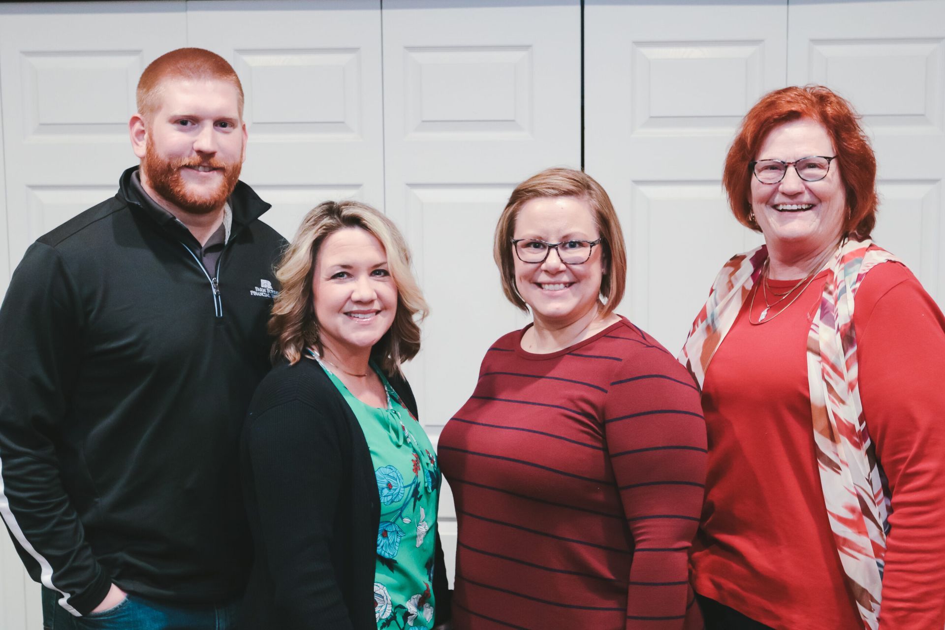 Four people smiling, posing together. Two women wearing glasses, one in a red shirt, one with a floral top. Two 