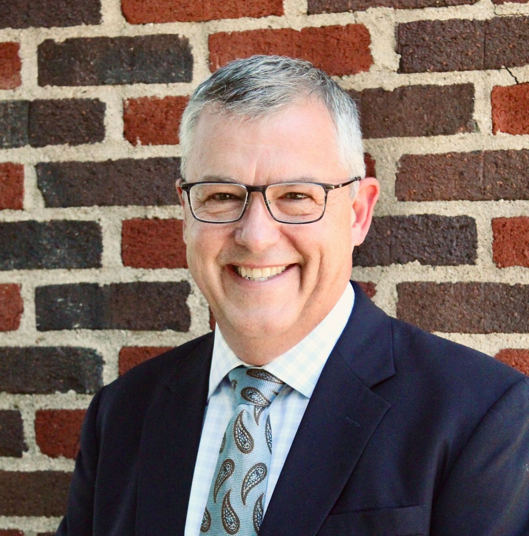 A man in a suit and tie is smiling in front of a brick wall.