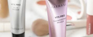 Primer plano de un tubo de base iluminadora Farmasi VFX PRO de color rosa con elementos de maquillaje difuminados en el fondo.