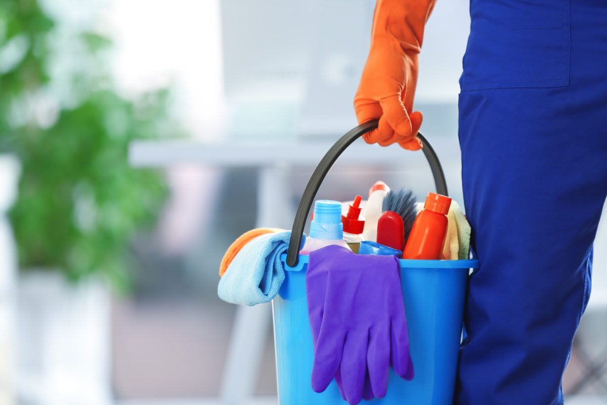 Housekeeper wearing blue coveralls, and orange gloves holding a light blue bucket full of cleaning supplies. 