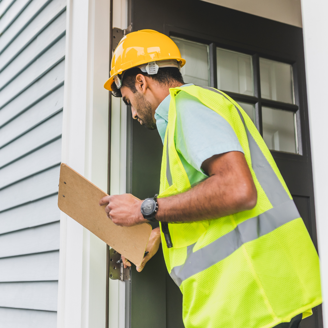 House inspector wearing a high-vis jacket and a hard hat holding a clip-board inspecting an outdoor door frame.