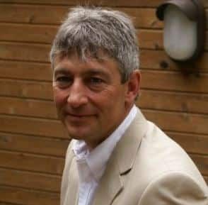 Man with graying hair, wearing a beige blazer and white shirt, smiles in front of a wood-paneled wall