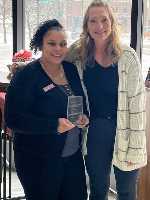 Two women standing indoors, one holding an award. The woman on the left is smiling, wearing black. 