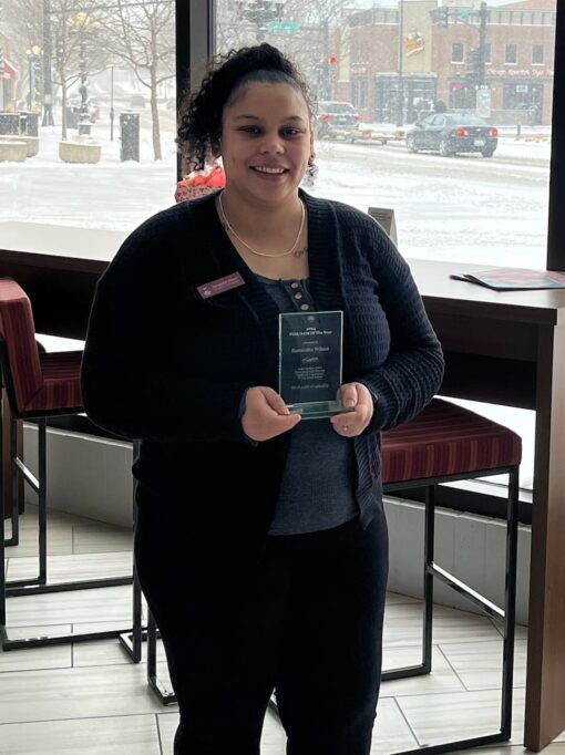 Woman holding an award, smiling. She's in a black cardigan and pants, standing near a window in a restaurant setting.