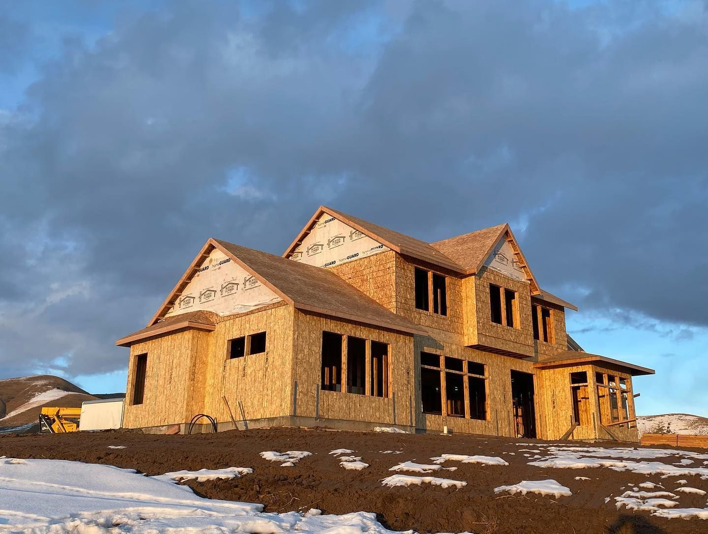 House under construction on a snowy hill under a cloudy sky. Wooden frame visible.