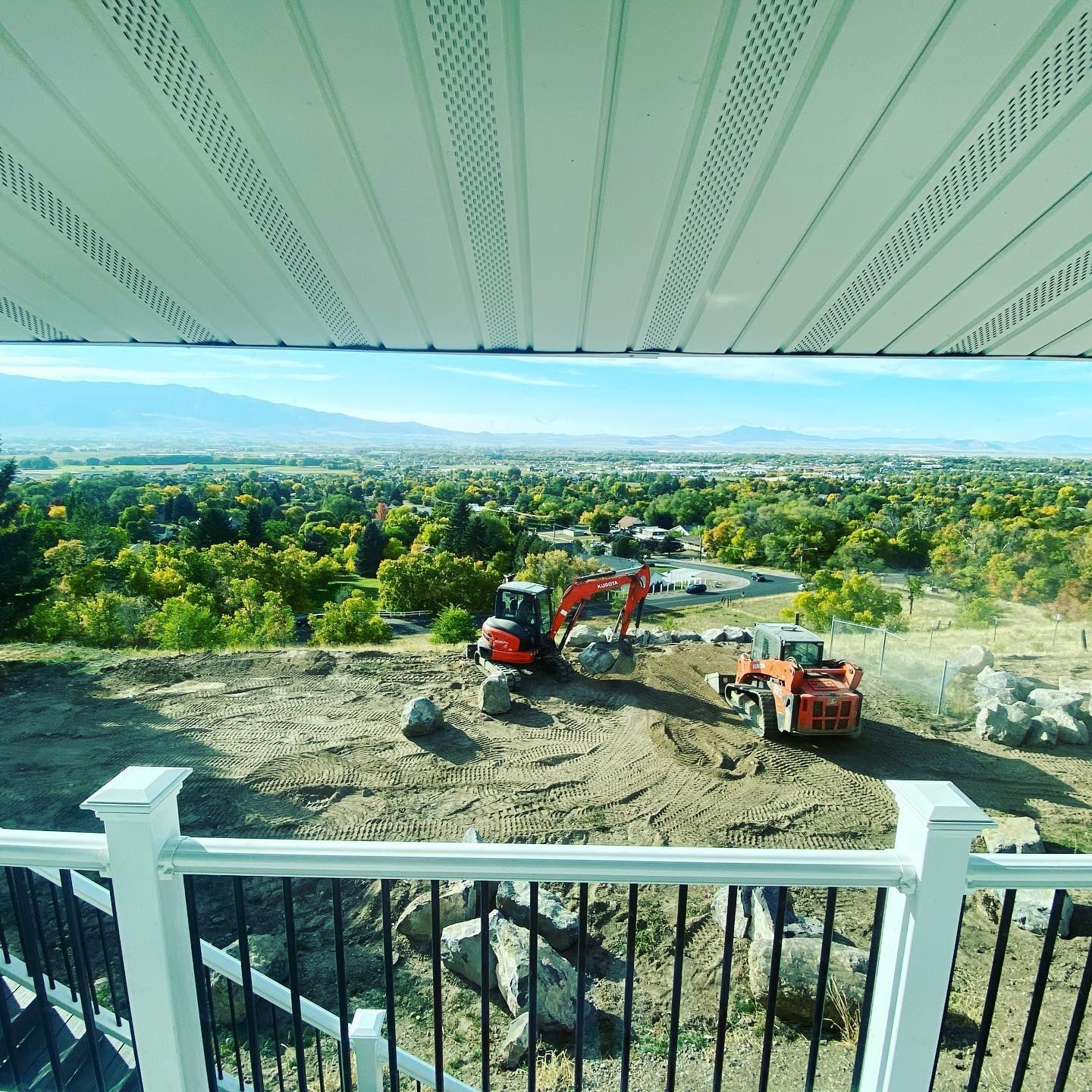 Construction site with excavators on a hillside, overlooking a valley with trees and a mountain range, viewed from a balcony.