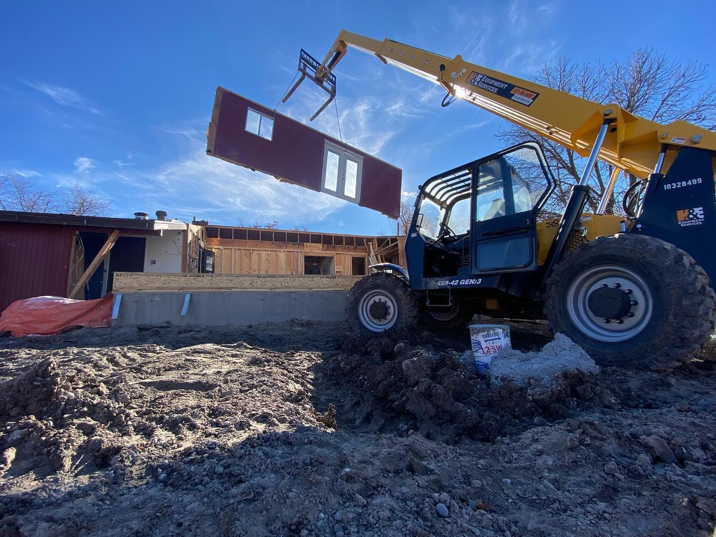 A telehandler lifts a red wall panel with a door and windows at a construction site.