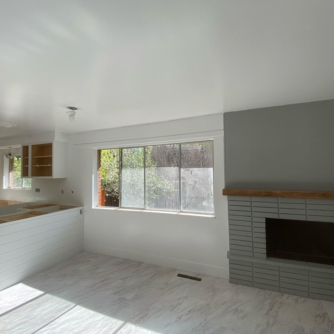 Kitchen and living area with gray fireplace and a window, white cabinets, and light gray walls and flooring.