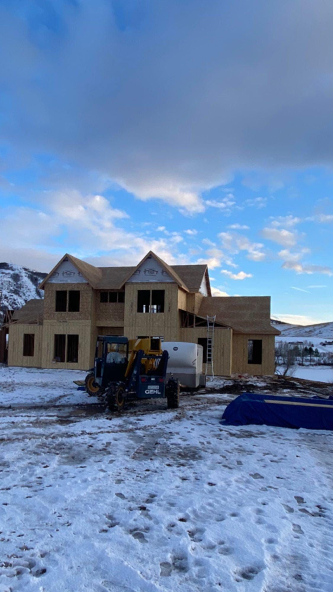 Wood-framed house under construction on a snowy lot; machinery and cloudy sky in background.